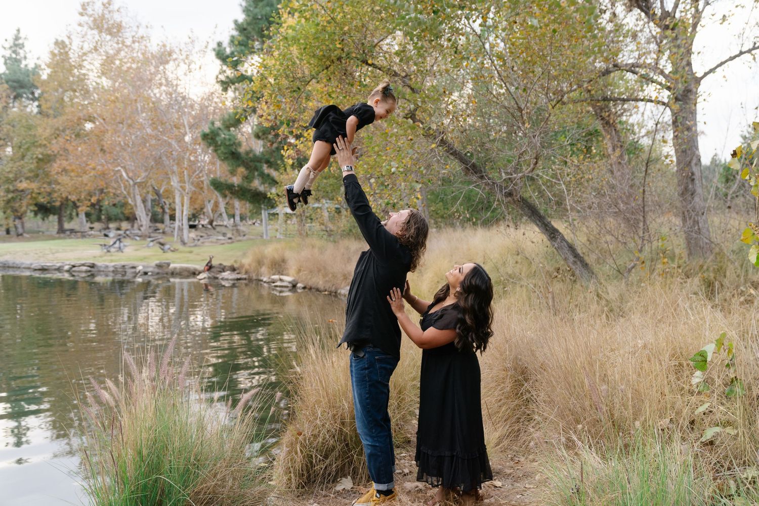 Parents joyfully lift child in air while standing by lakeside during outdoor photoshoot.