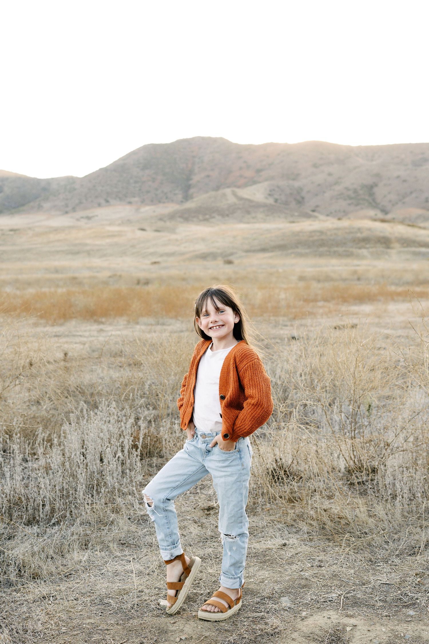A smiling girl in an orange cardigan, white top, and ripped jeans poses in a dry golden field with hills behind her.
