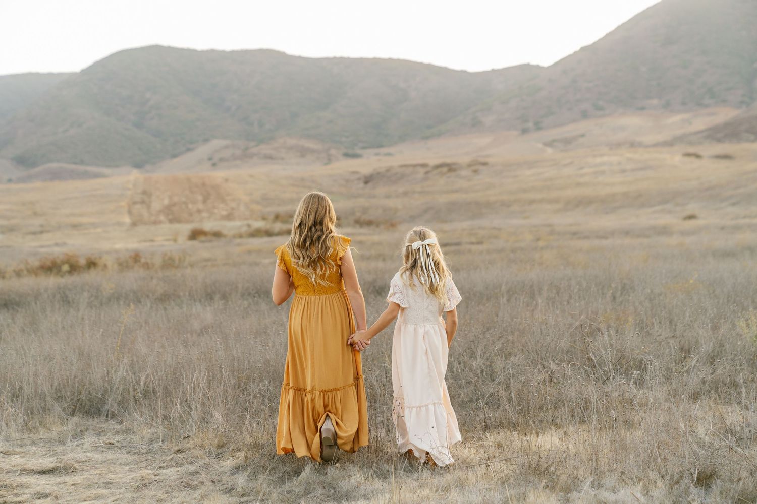 Two females in boho dresses hold hands in golden field with mountain backdrop.
