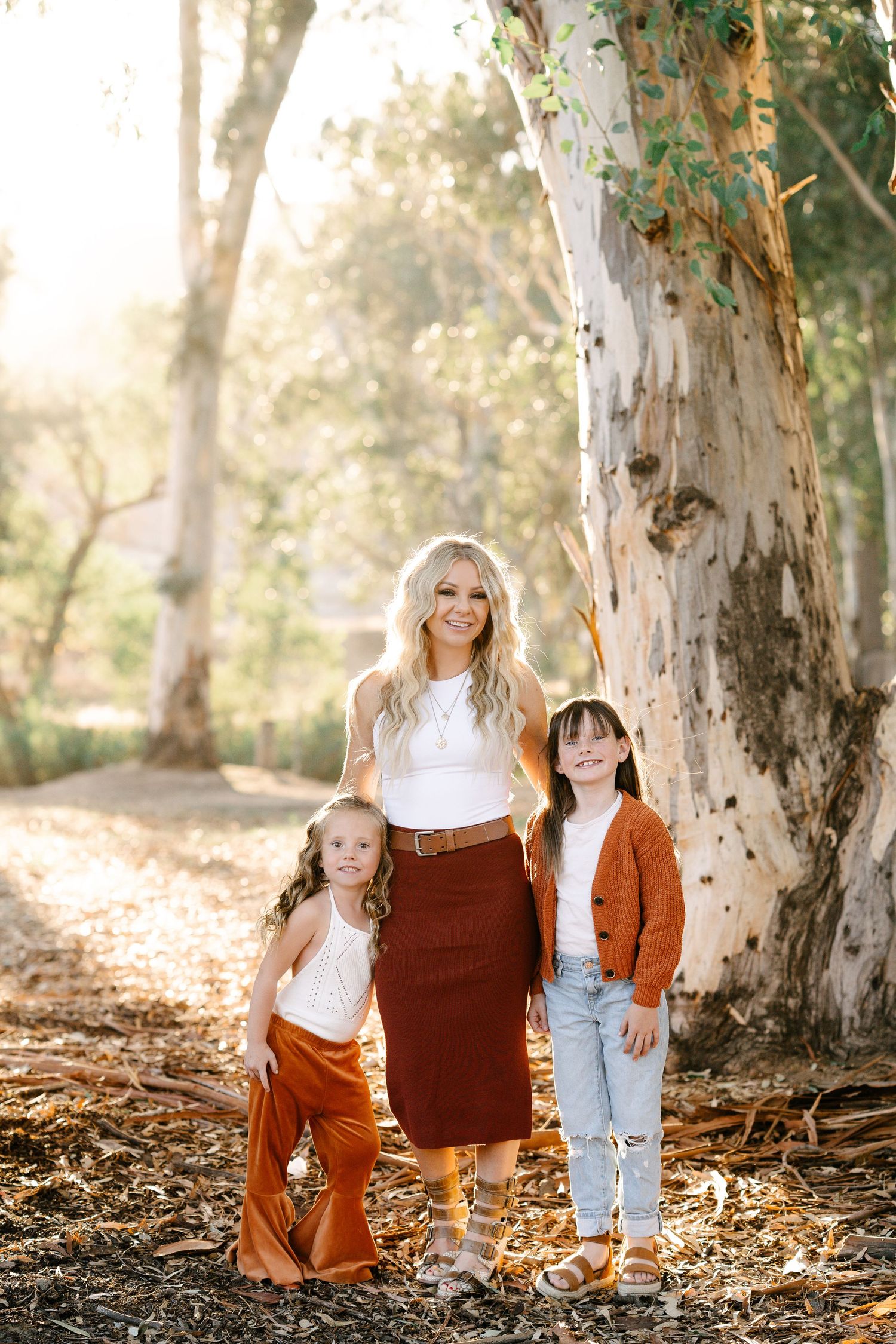 Woman and two girls pose outdoors near large tree in warm autumn tones, sunlight filtering through eucalyptus trees.