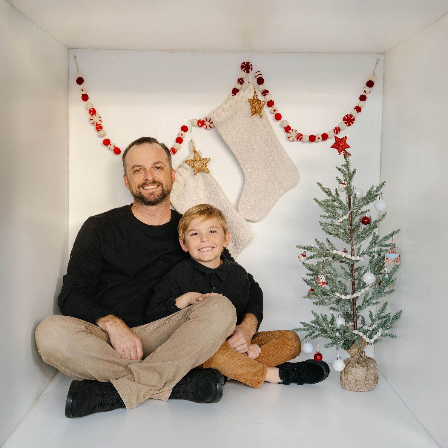 Father and child sitting together in front of Christmas decorations, stockings, garland, and a small decorated tree.