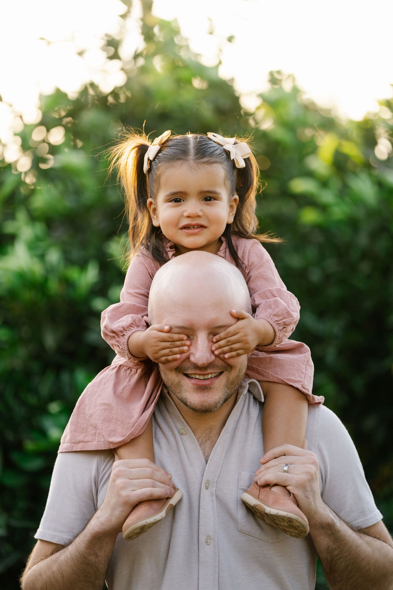 Playful moment as a toddler in pink covers someone's eyes while sitting on their shoulders.