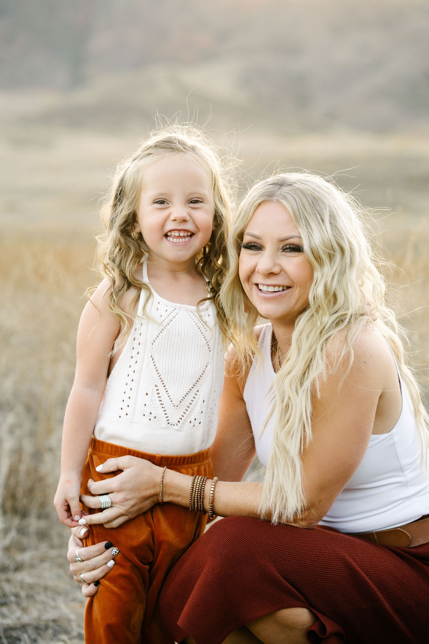 A smiling blonde mother crouches beside her curly-haired daughter in a golden field at sunset.