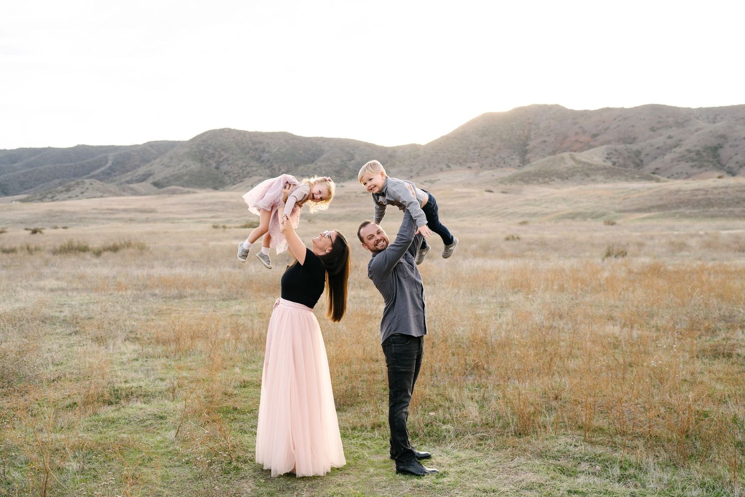 A joyful family lifts two young children in a scenic open field with rolling hills in the background.