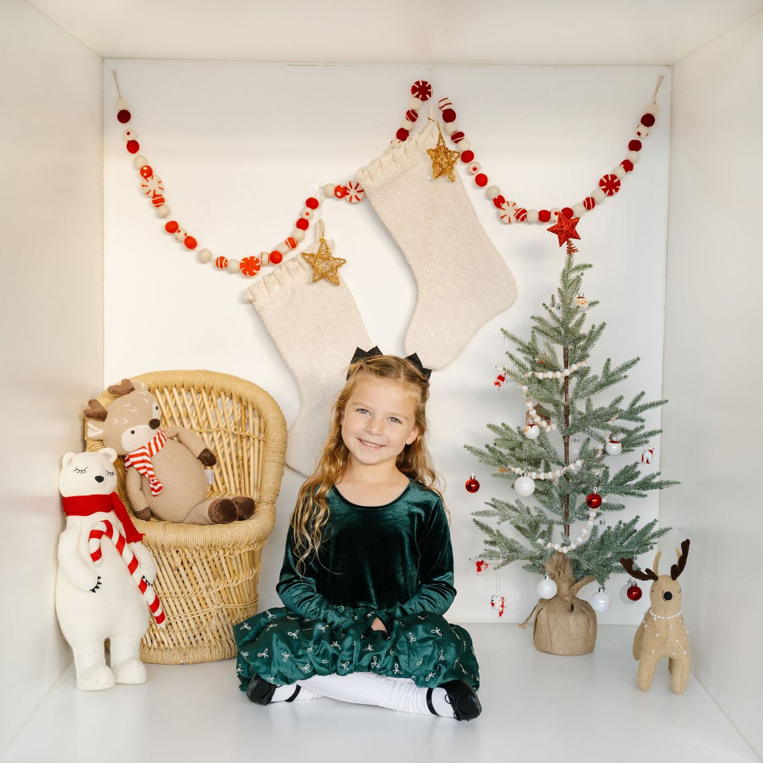 Young girl in green velvet dress sits before Christmas tree, stockings, and holiday garland decor.
