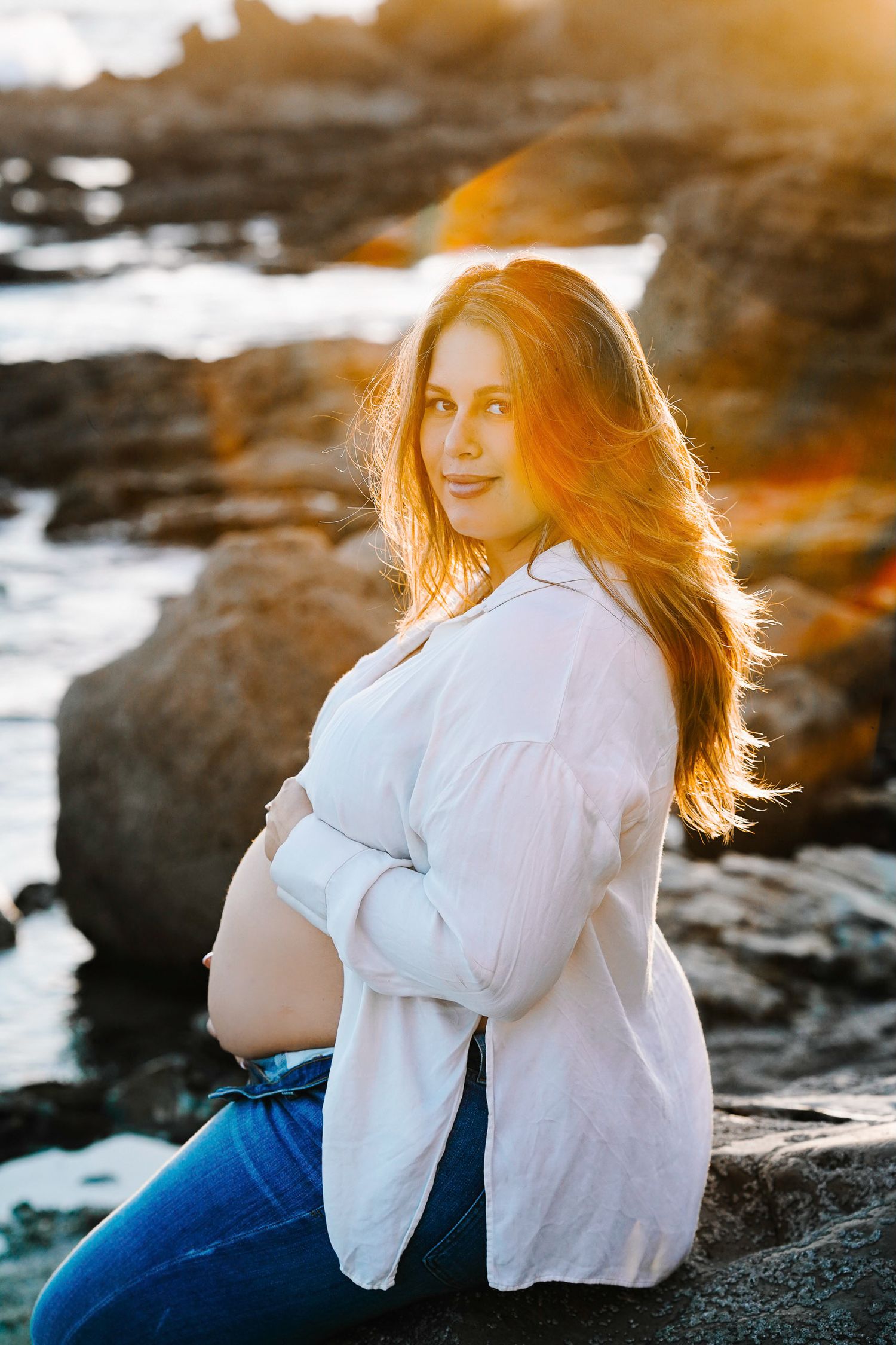 Maternity photo of expecting mother sitting on coastal rocks during golden hour sunset.