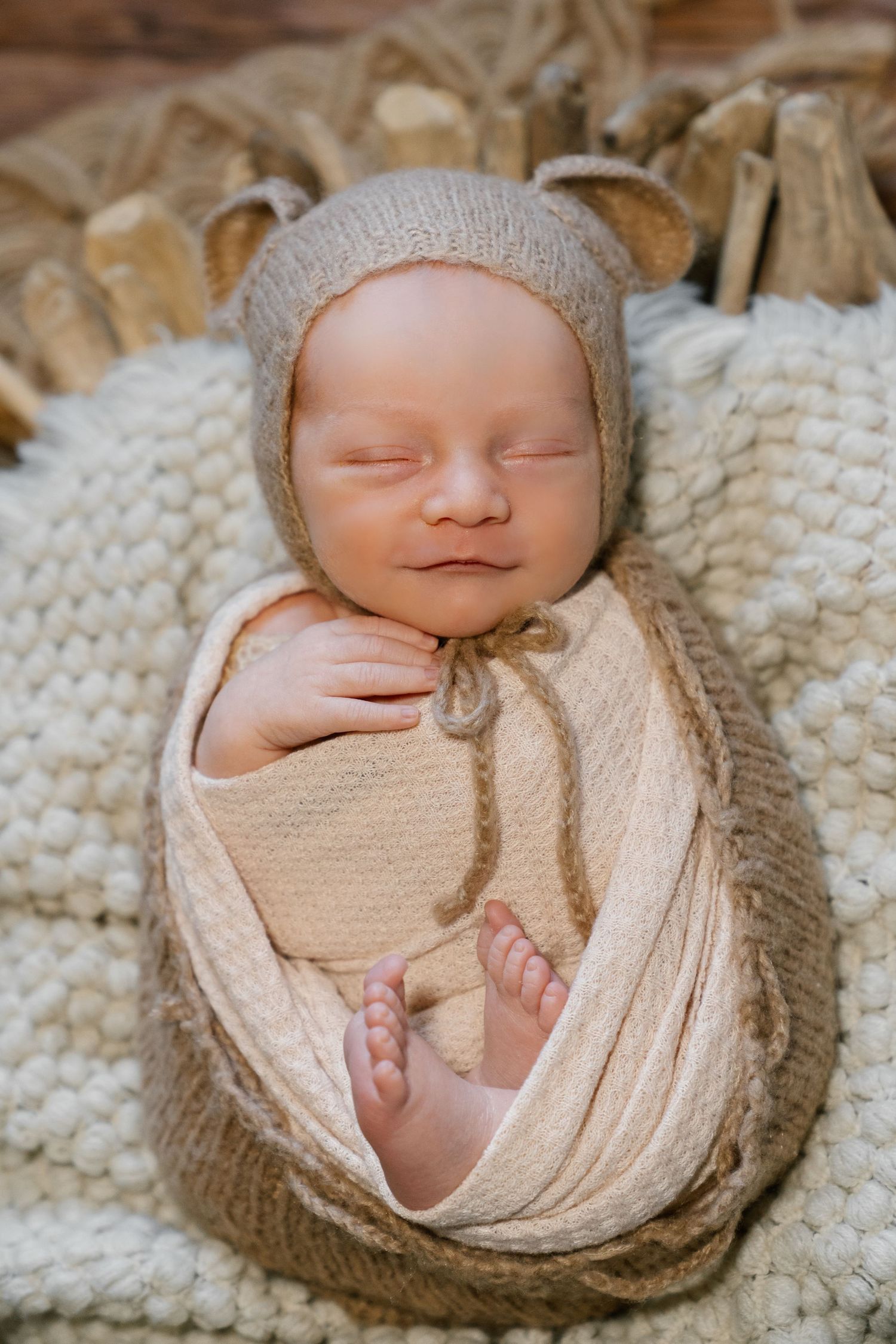 Newborn baby wrapped in cream knit swaddle wearing matching bonnet on textured blanket.