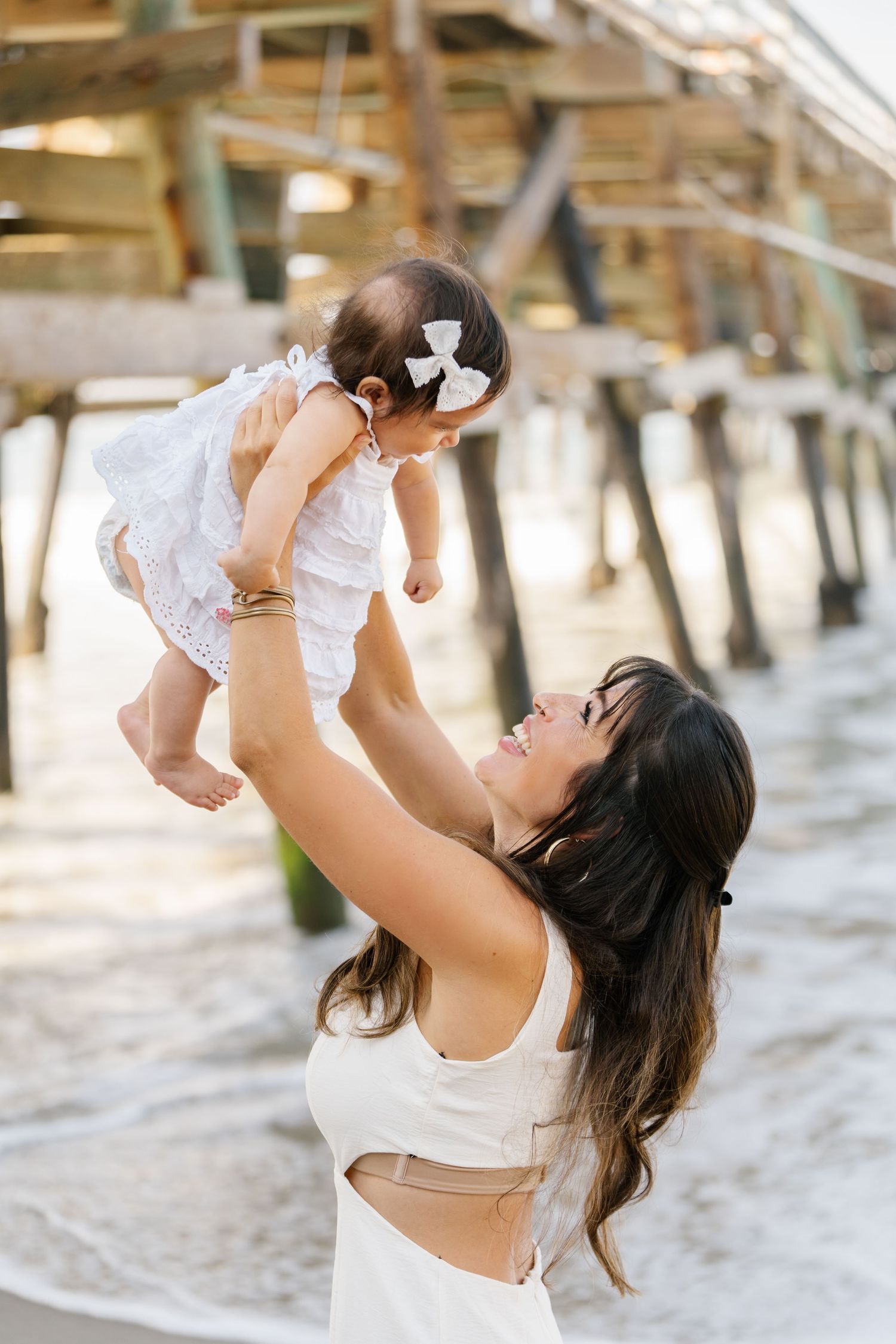 Mother lifting baby in white dress by beach pier.