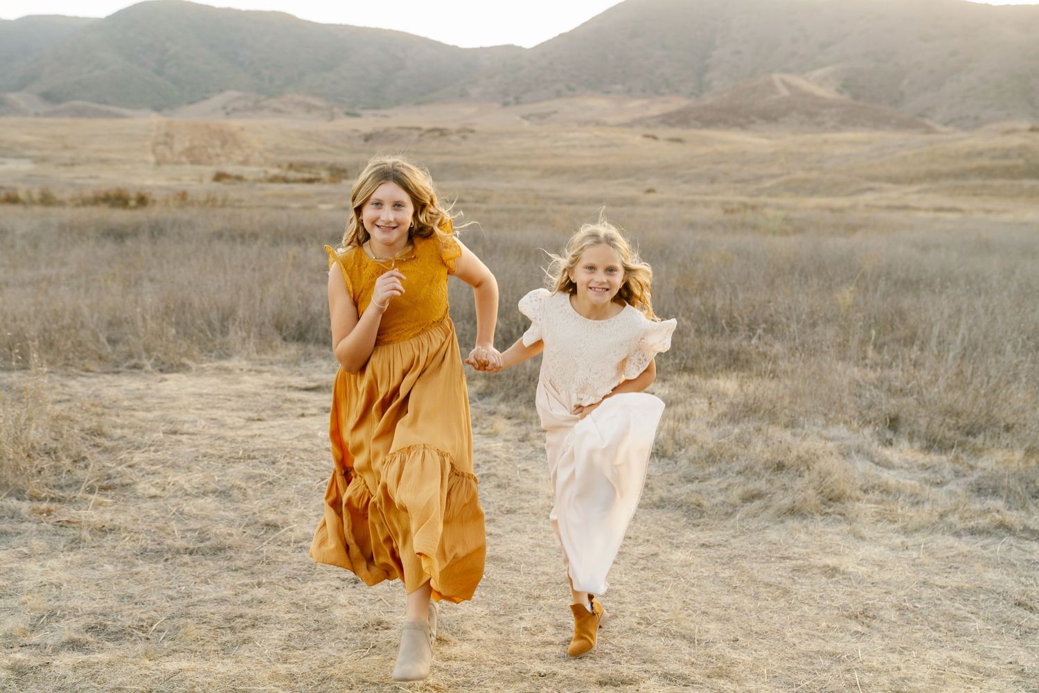 Two girls in flowing dresses run joyfully through a golden field with rolling hills in the background.