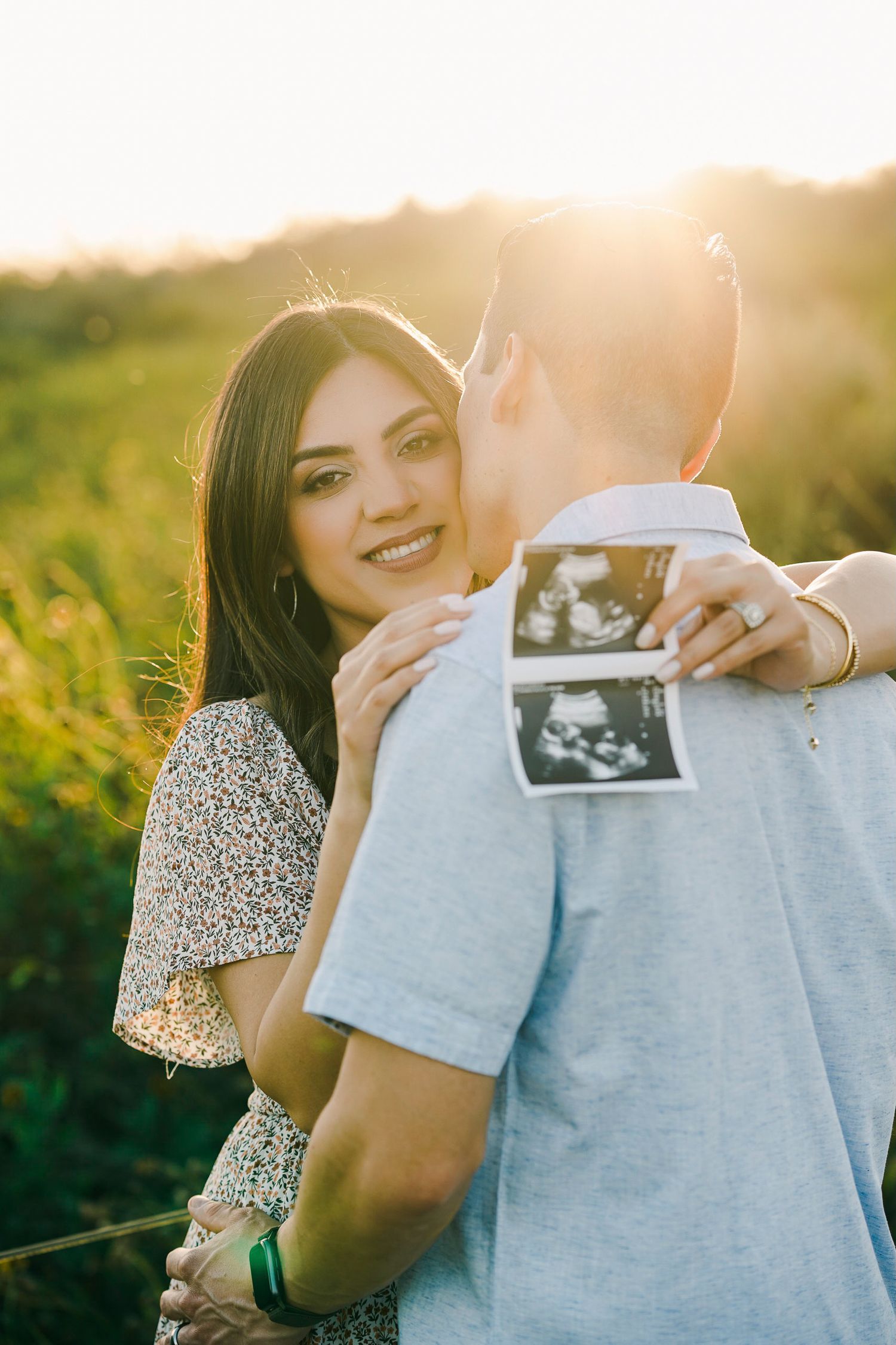 Couple holds ultrasound photo while embracing in golden sunlit field during sunset pregnancy announcement.