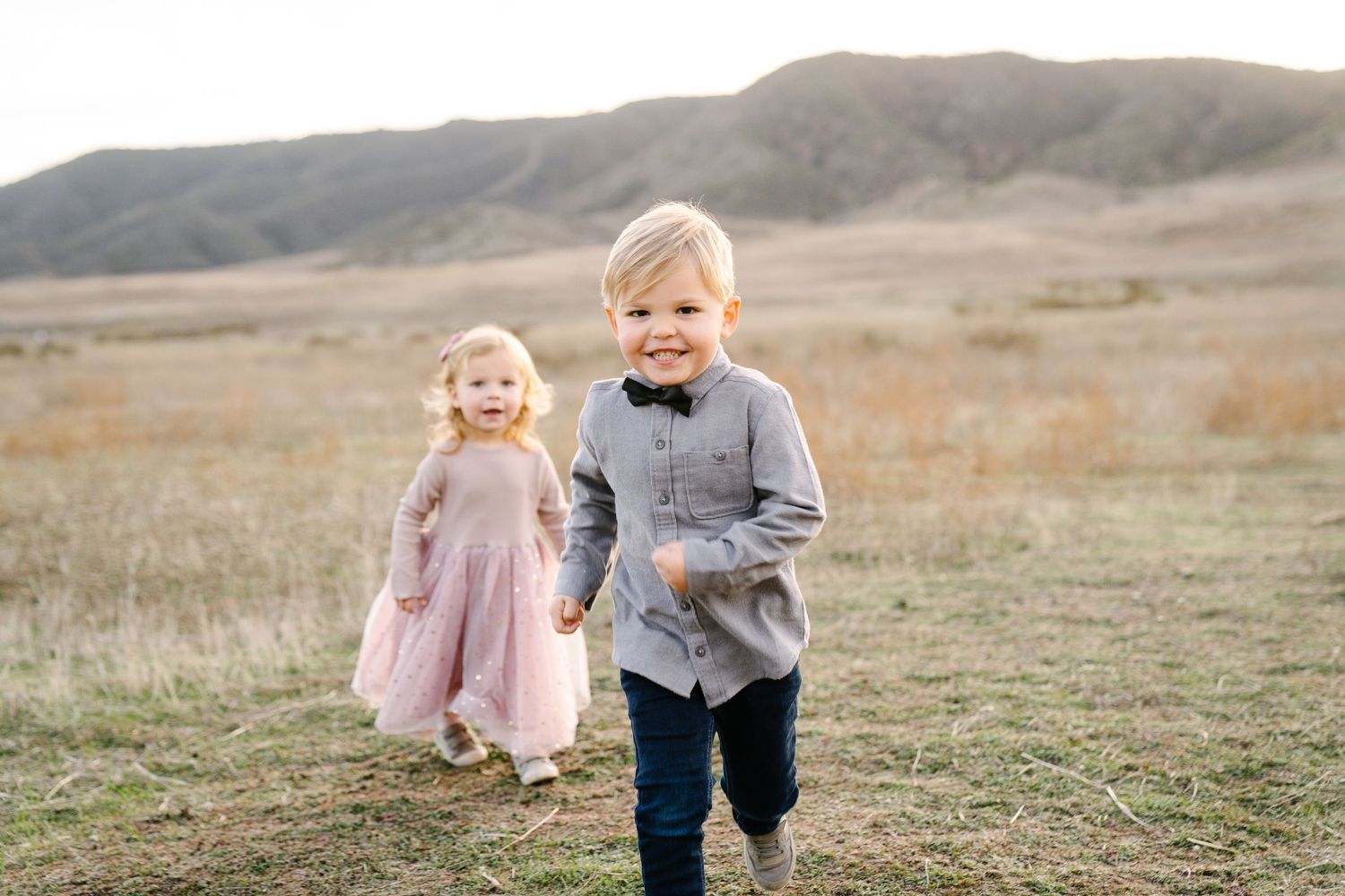 Two young children running playfully through a grassy field with mountains in the background.