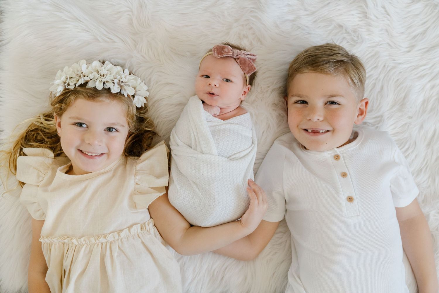 Three children in white outfits pose together on light bedding.