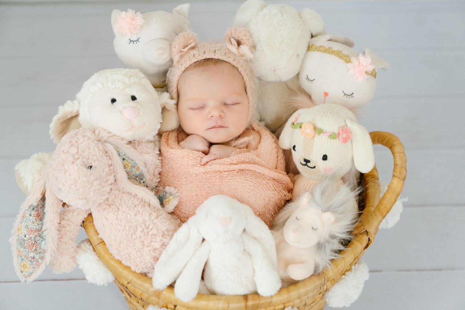 A sleeping baby nestled in a basket surrounded by plush bunny and lamb toys.