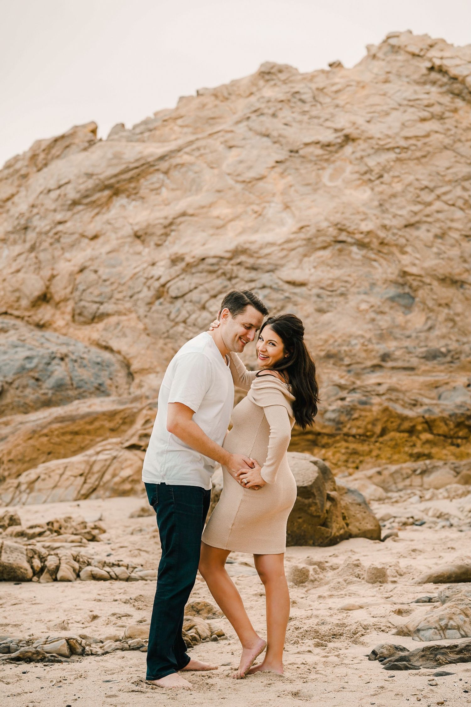 Loving couple embraces on a rocky beach during a maternity photoshoot.