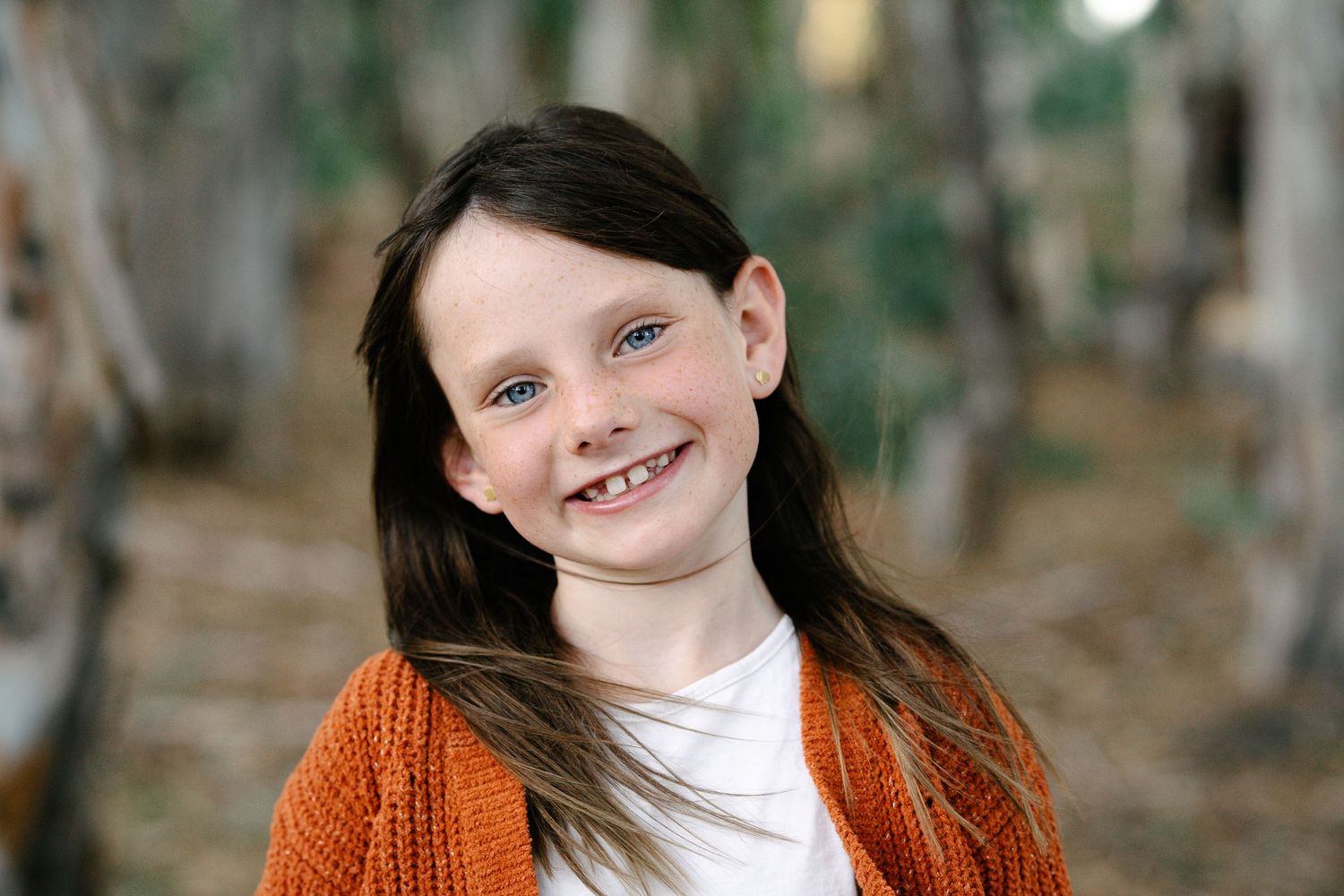 Smiling young girl with long dark hair wearing an orange cardigan outdoors among trees.