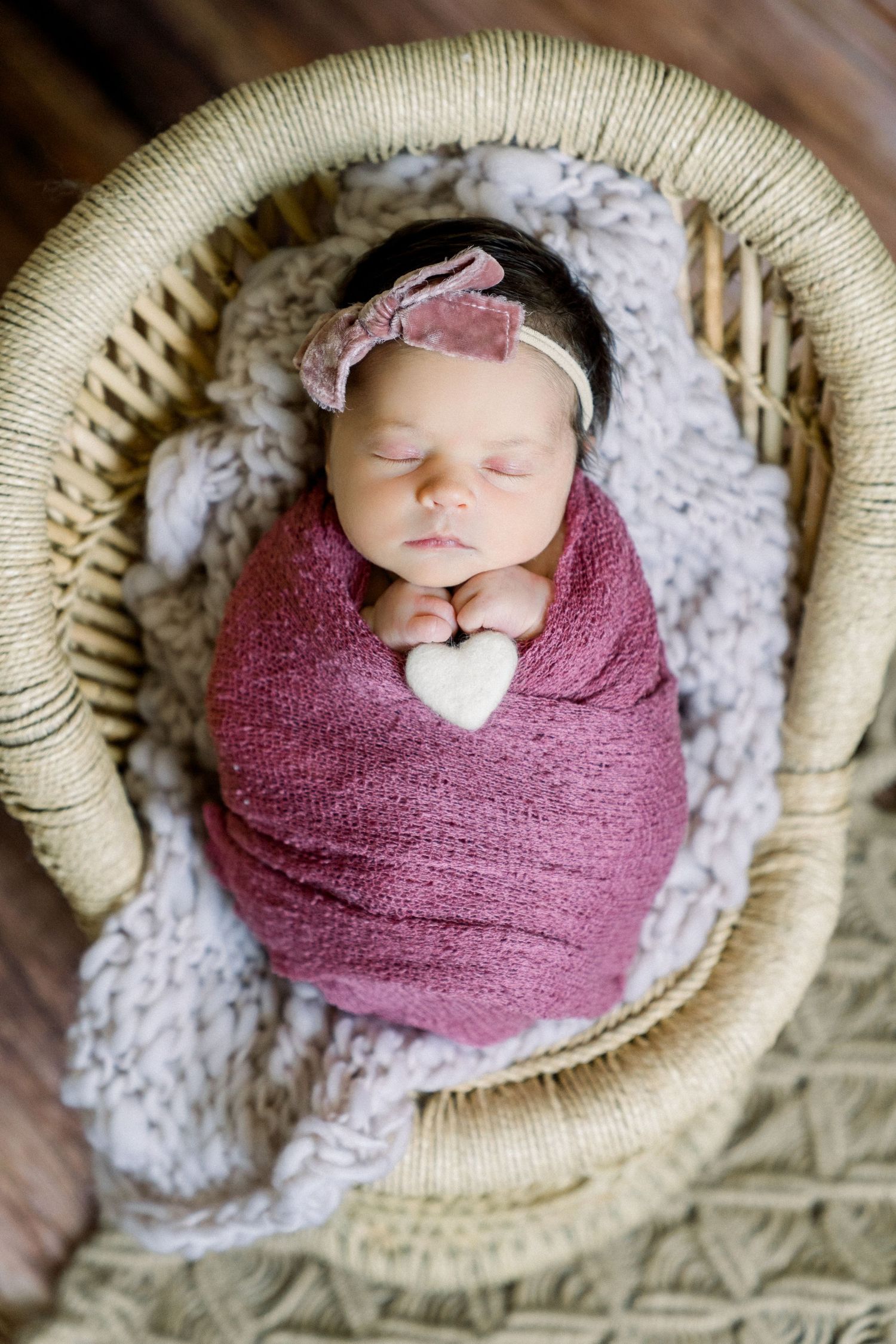 Peaceful sleeping baby swaddled in burgundy wrap with bow headband in round basket.