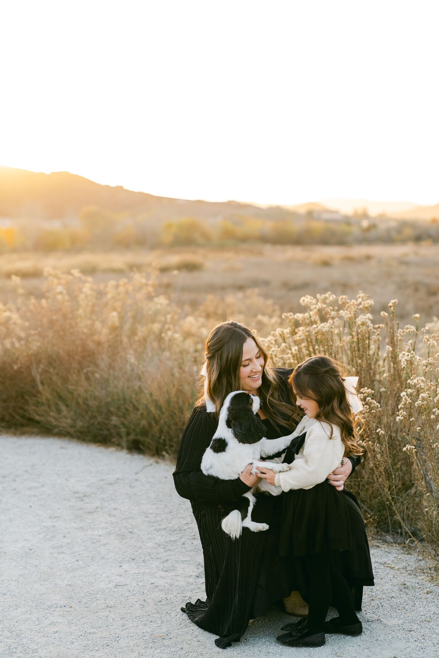 Two people in black dresses share a sweet moment with a black and white dog on a sunset trail.