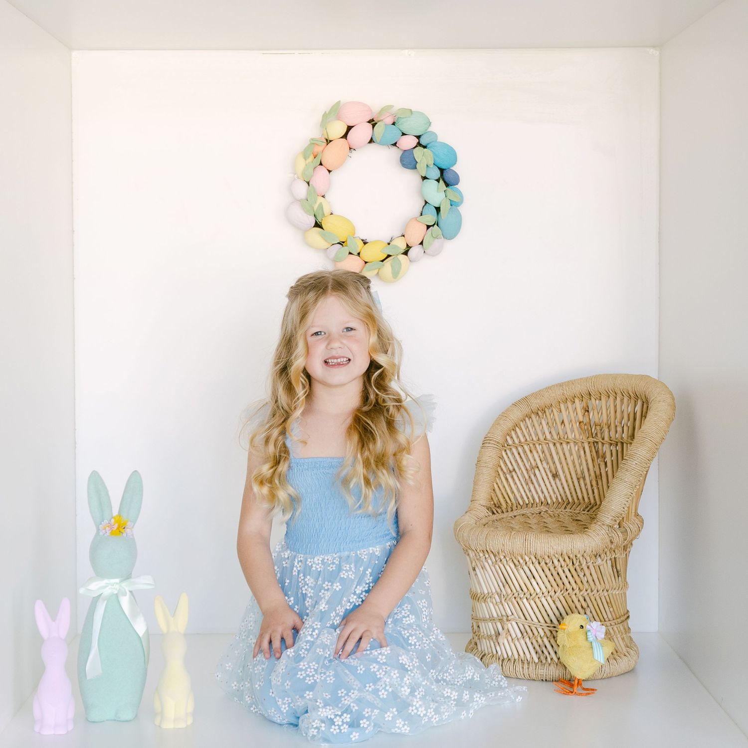 A pastel Easter scene with a floral wreath, wicker chair, decorative bunnies and soft spring decor against a white wall.