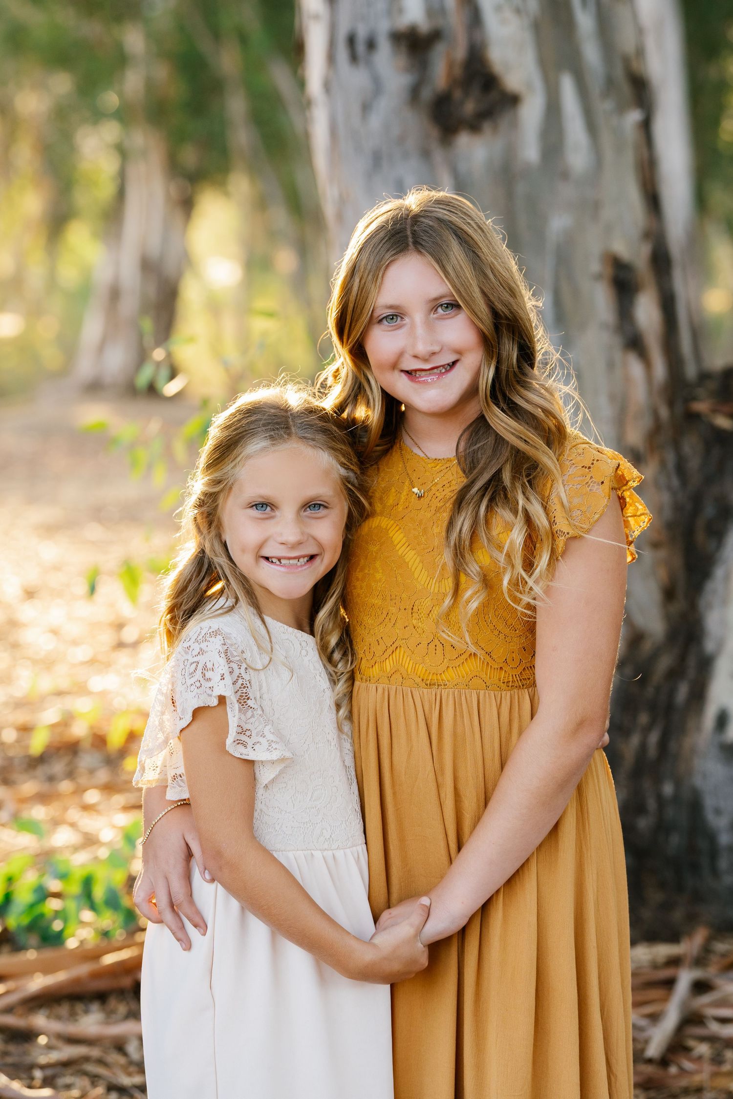Two smiling girls in white and mustard dresses pose together outdoors among trees in warm golden light.