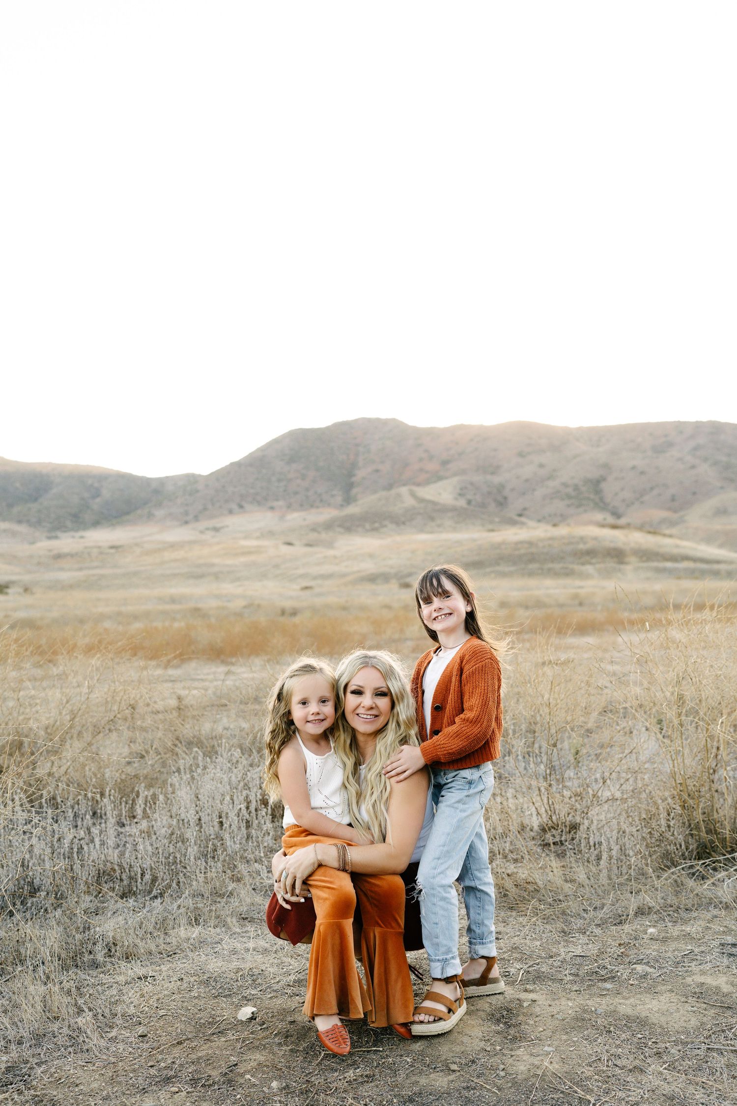 Mother and two daughters in coordinating rust orange outfits pose together in a golden hillside meadow.