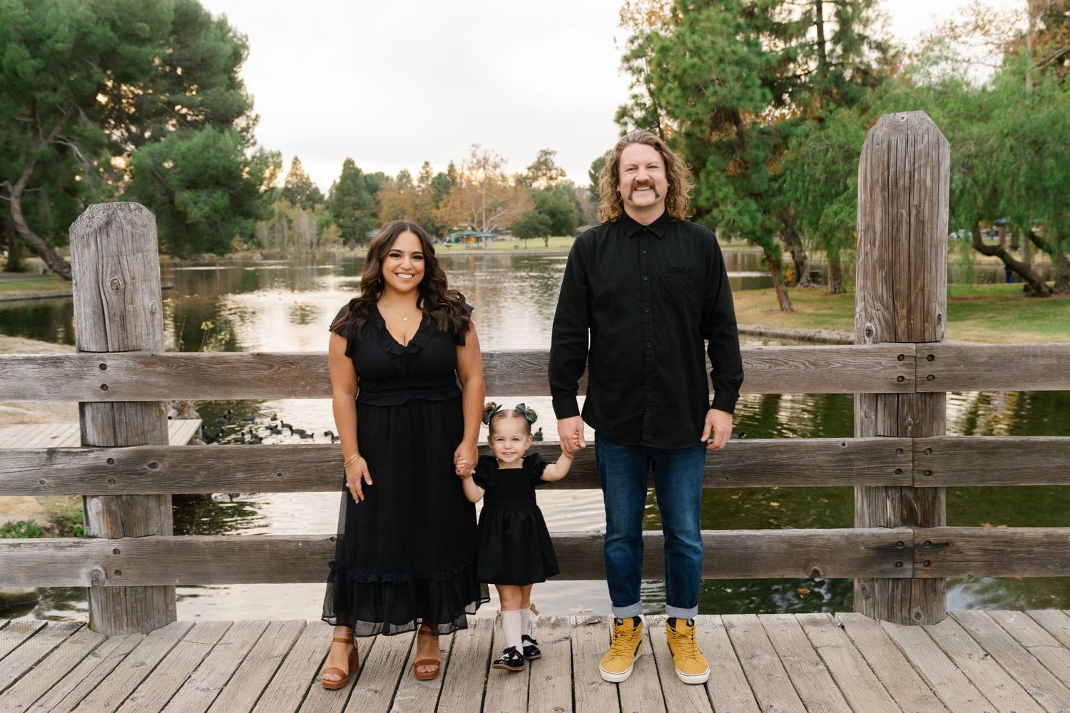 Family poses together on wooden bridge overlooking pond wearing coordinated black outfits.