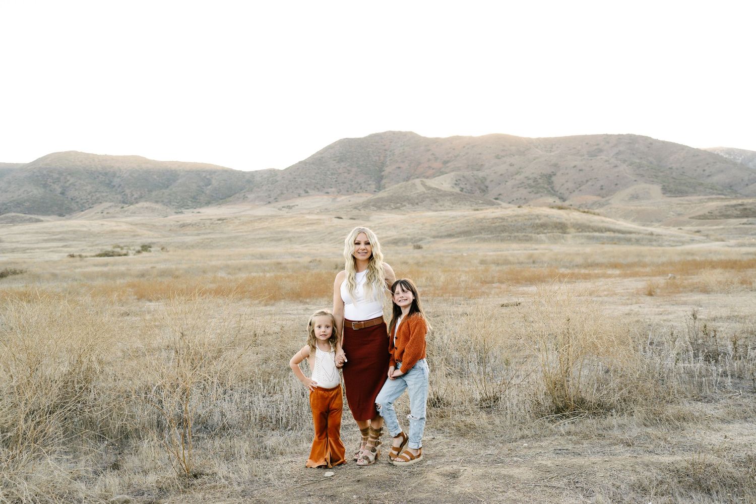 A woman and two children pose in a golden field with rolling hills in the background during a family photo session.