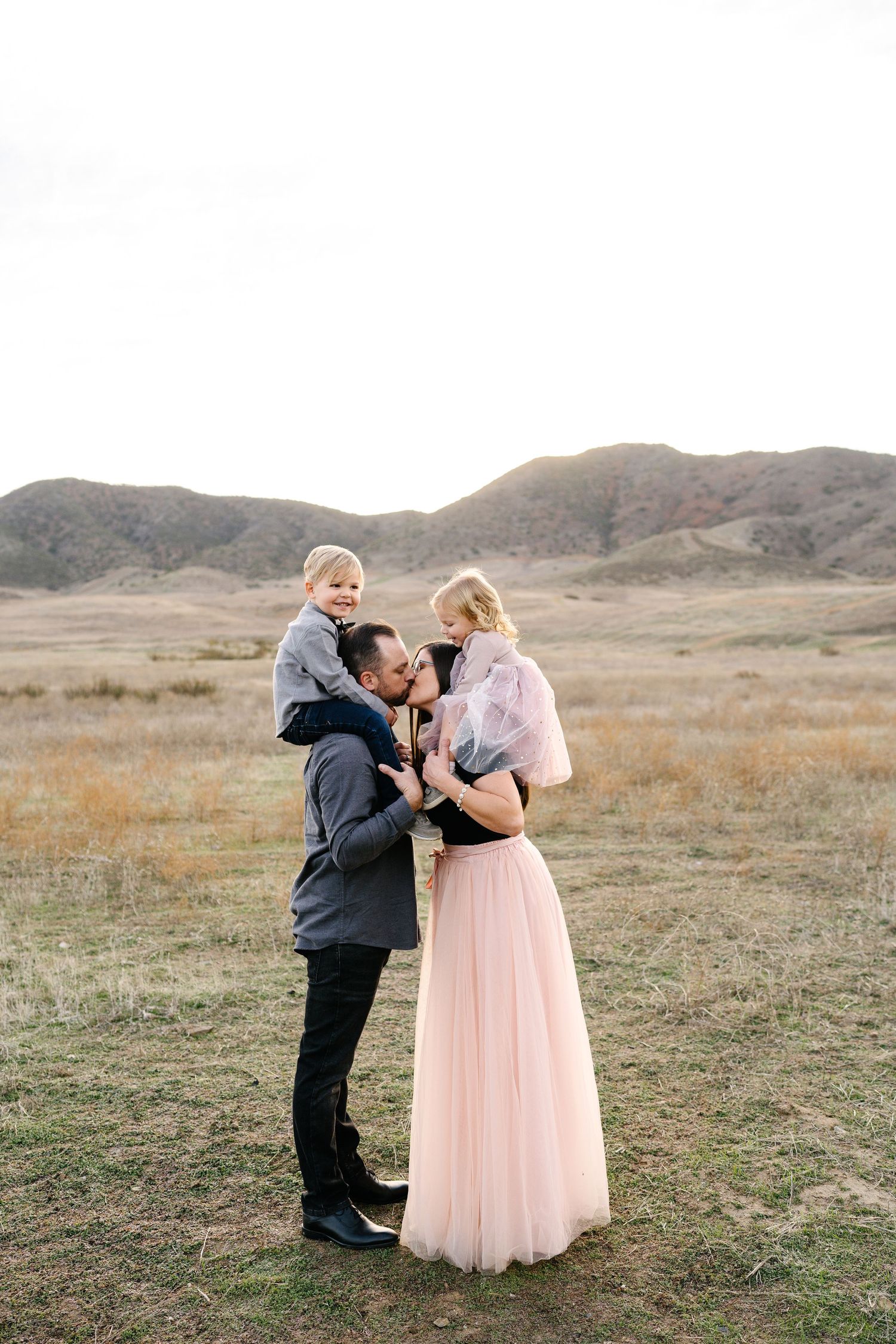 Family of four posing outdoors in a scenic field with rolling hills, father kissing mother while holding two young children.