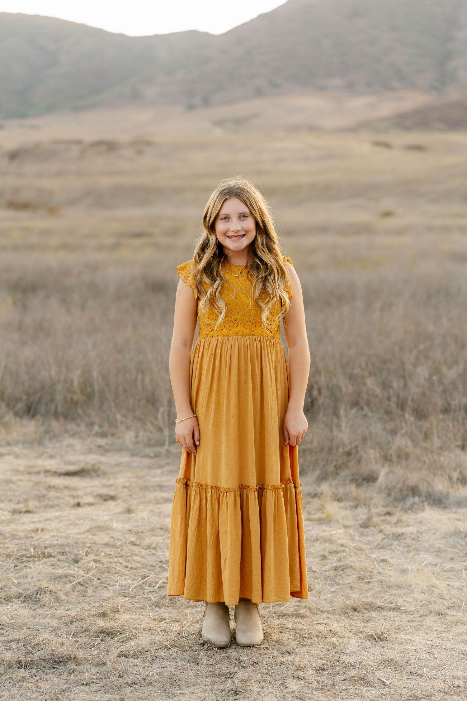 Young girl in mustard yellow maxi dress smiling outdoors in a dry grassy field.