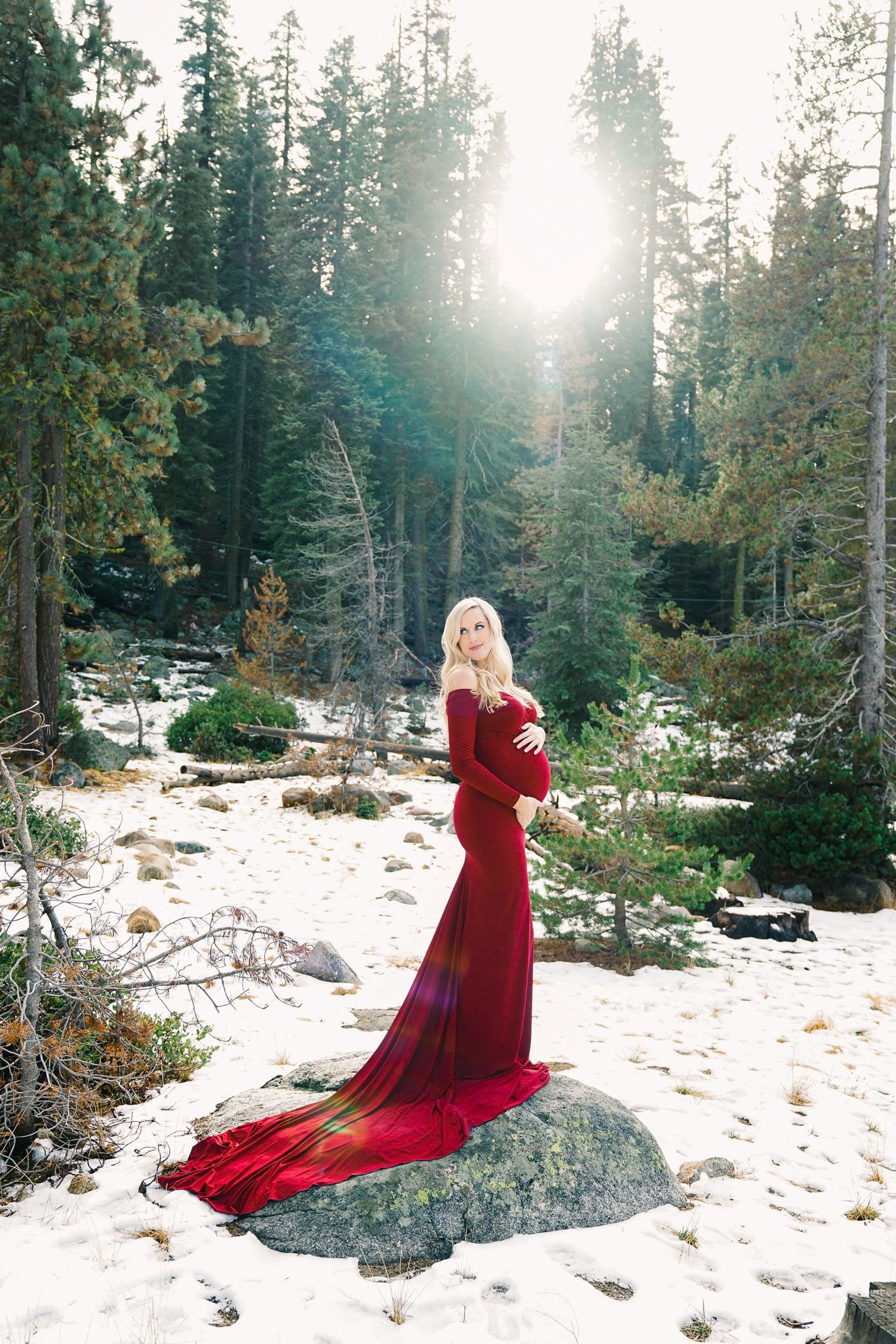 Dramatic maternity portrait in red gown against snowy forest landscape.