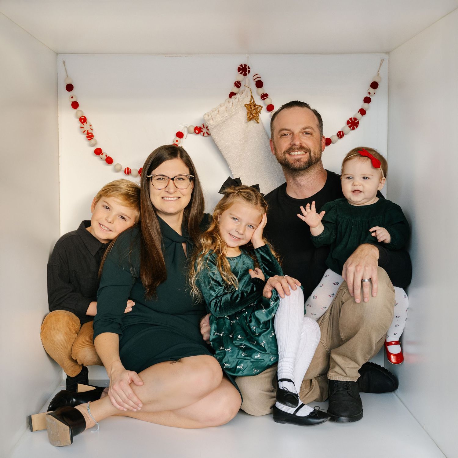 Family of five posing for a festive Christmas photo with holiday garland decorations in a white box setting.