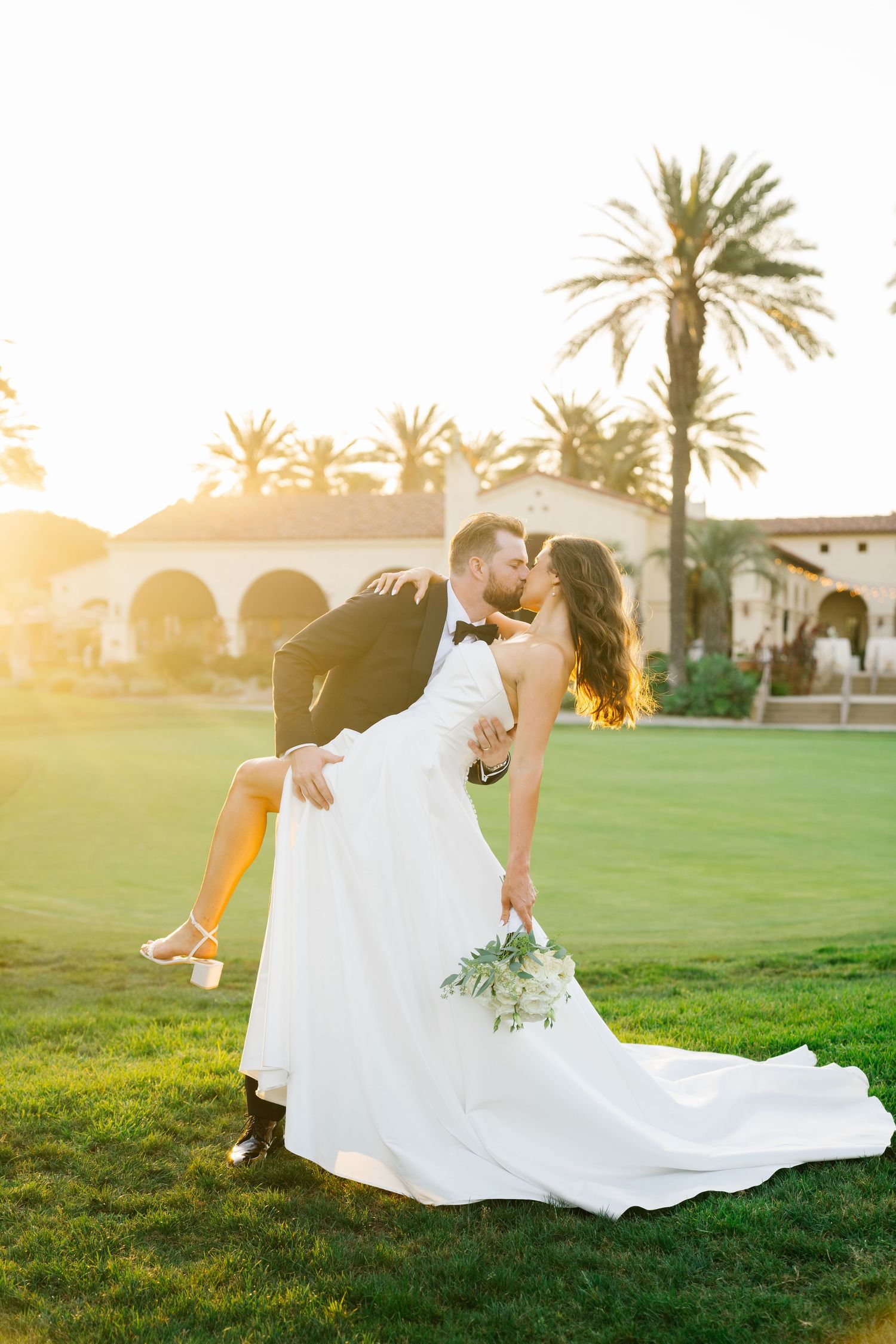 A romantic wedding photo captures a couple in a dramatic dip pose at sunset with palm trees in the background.
