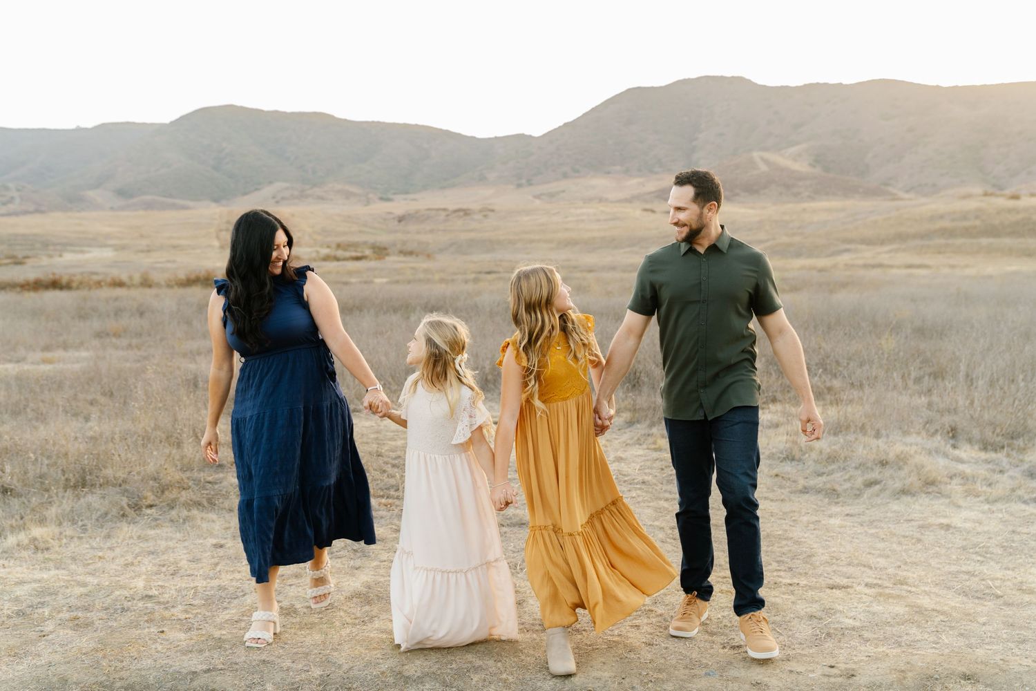 Family of four holding hands and walking through a golden field with scenic hills in the background.