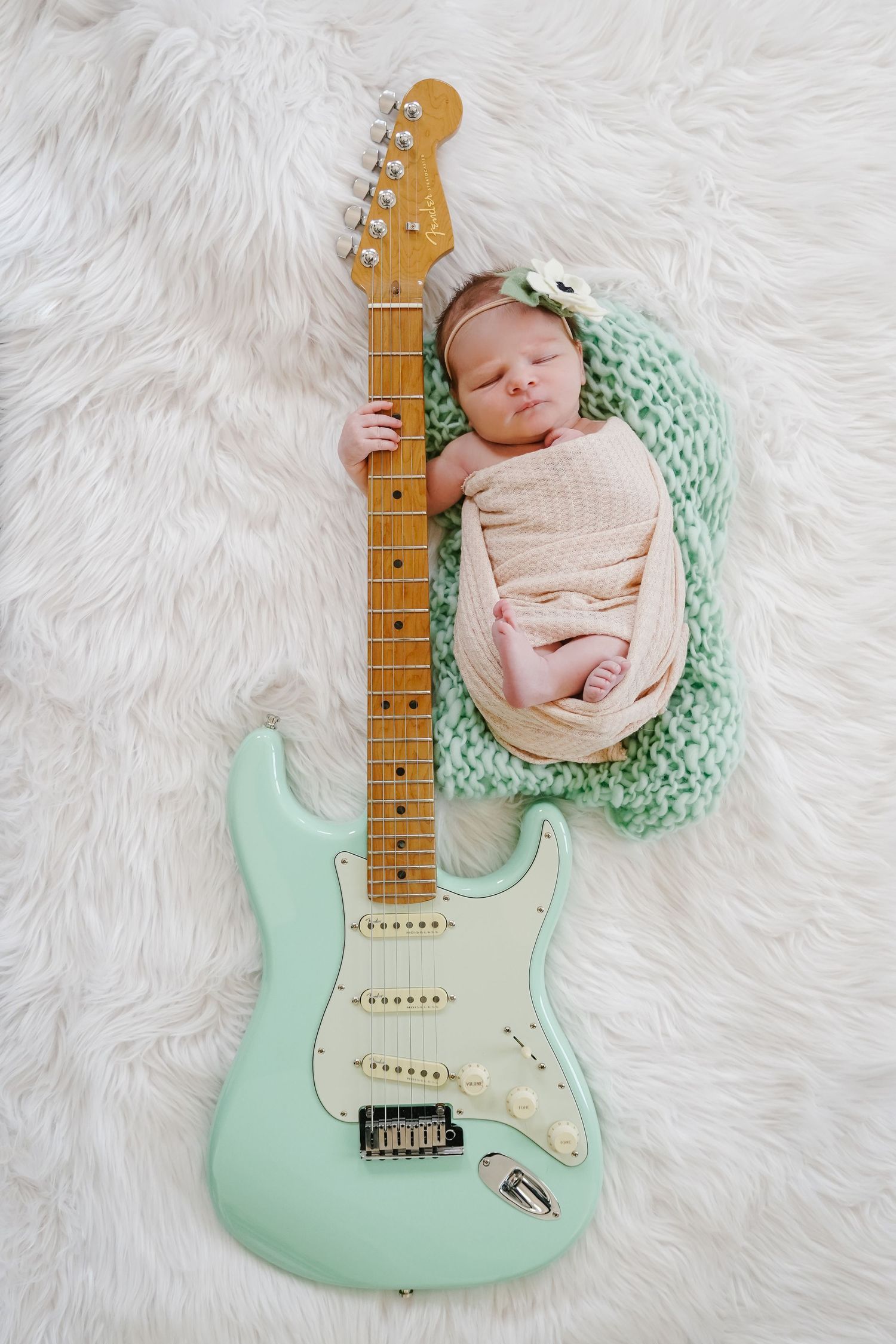 Newborn baby sleeping next to mint green electric guitar on white fur.