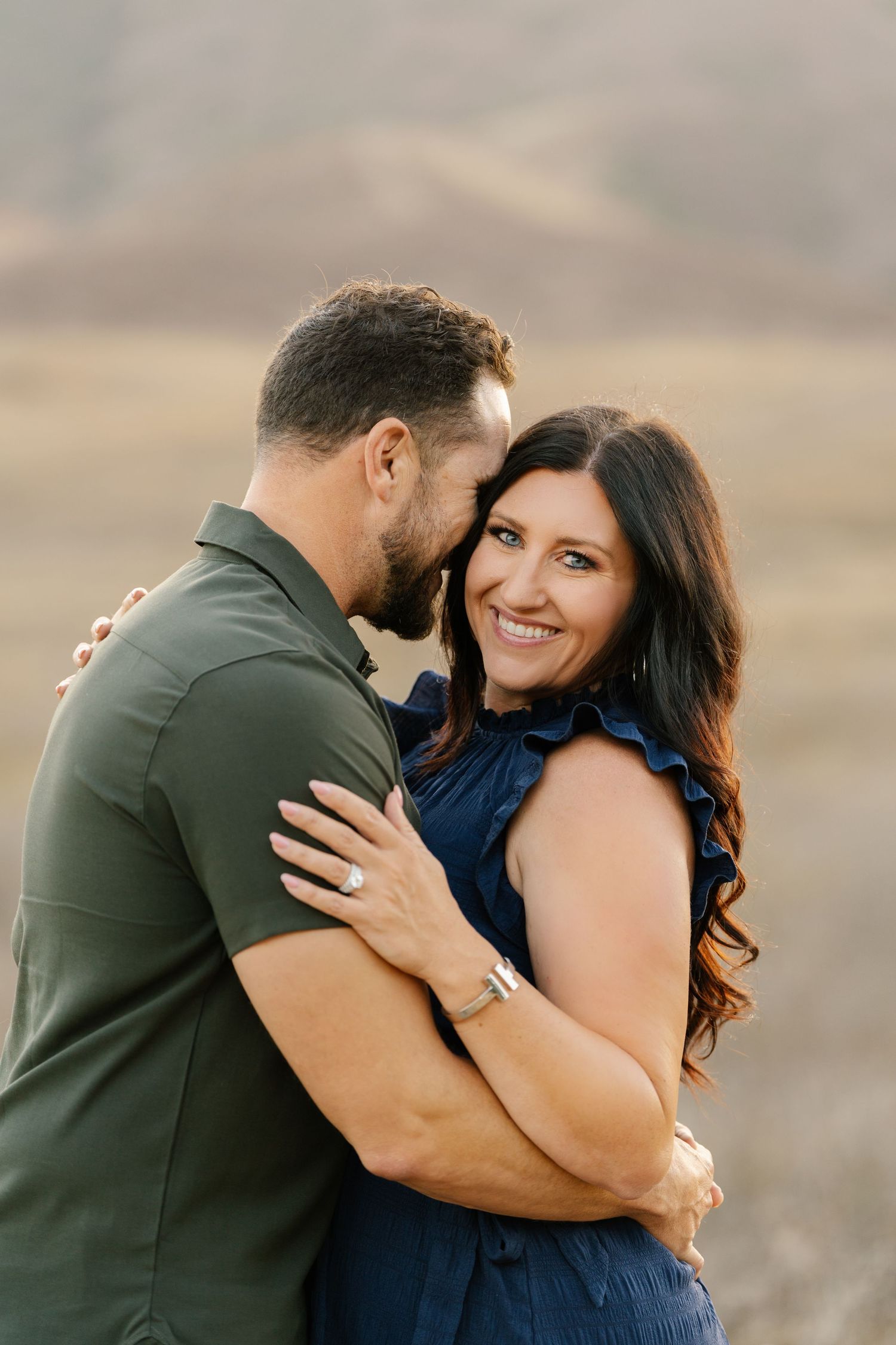 A smiling couple embraces outdoors, the woman in a blue dress and man in an olive shirt, in a scenic field.