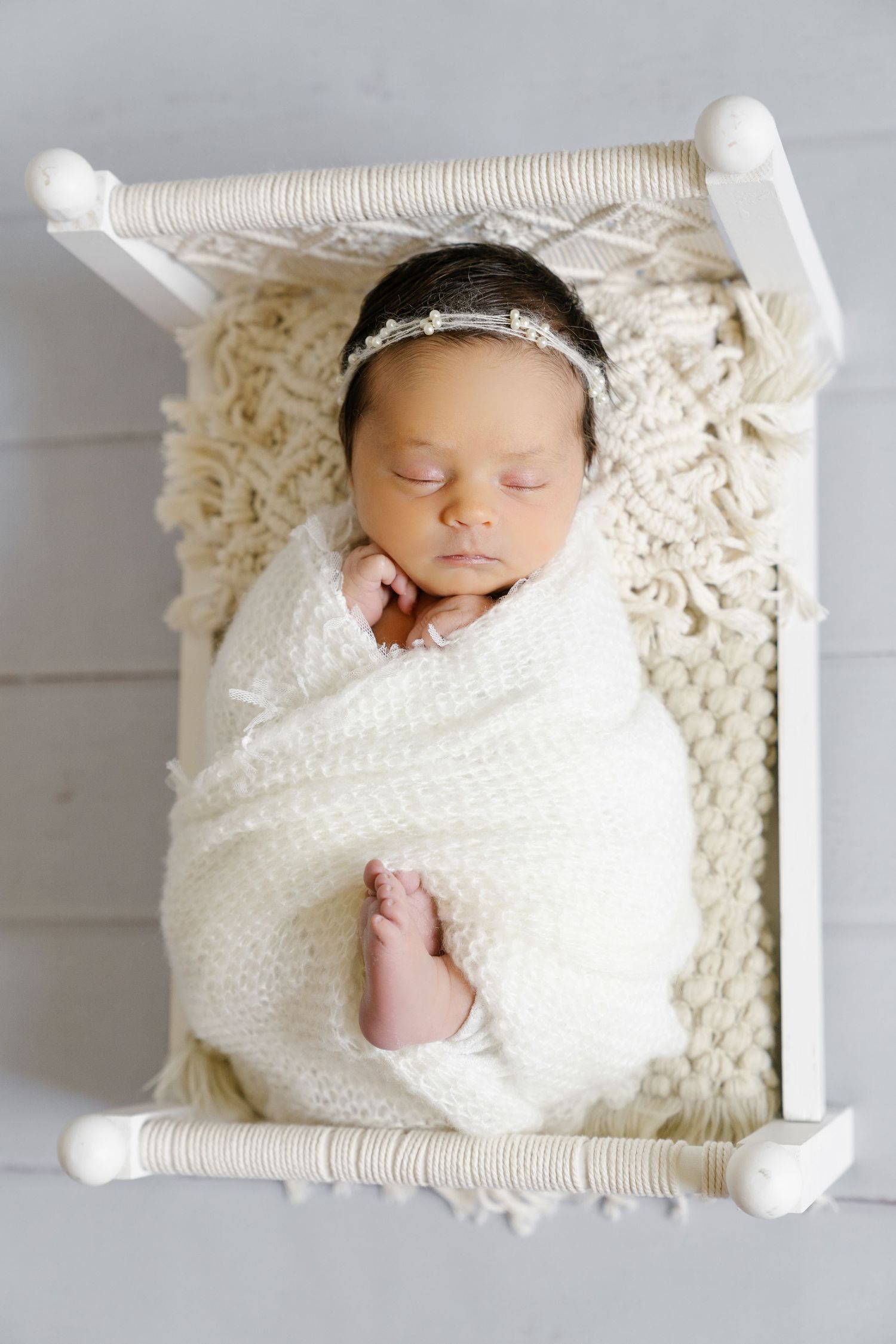 Peaceful sleeping newborn baby wrapped in white blanket on small white bed with lace headband.