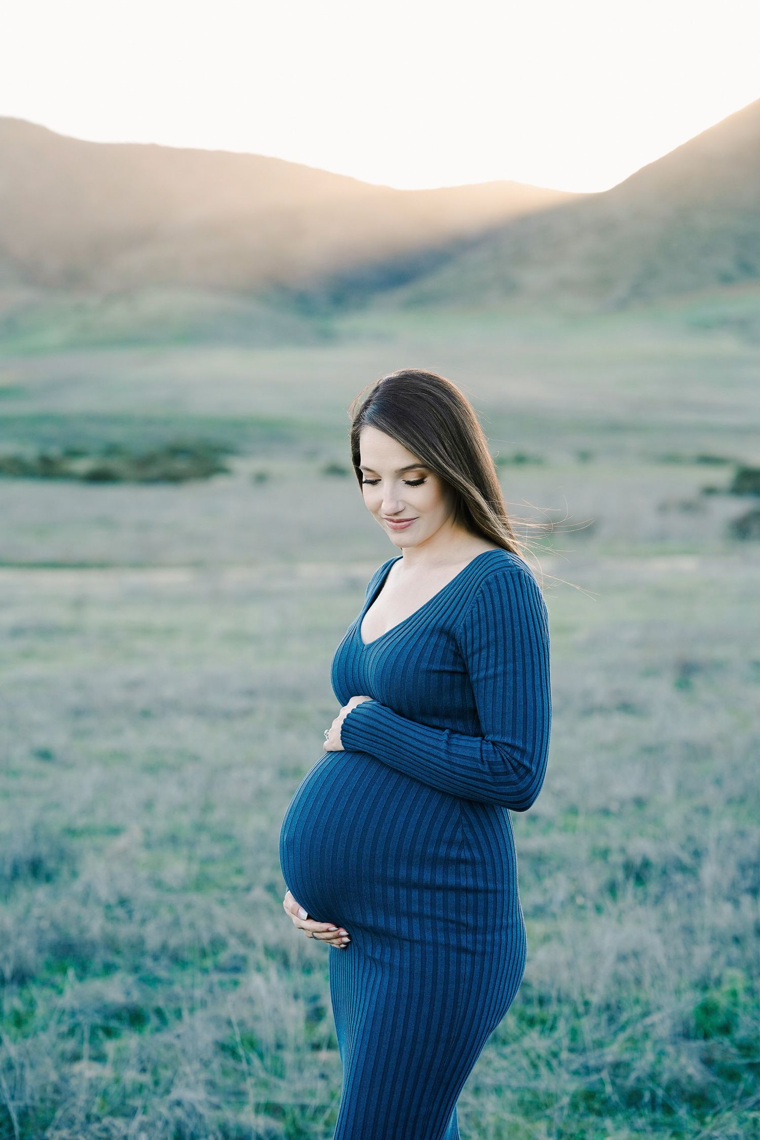 Expecting mother in blue knit dress poses in mountain meadow during golden hour.