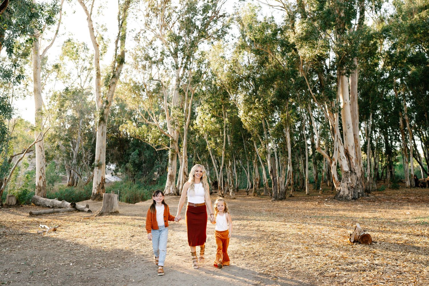 A mother walks with two young children through a sunlit eucalyptus grove on a dry dirt path.