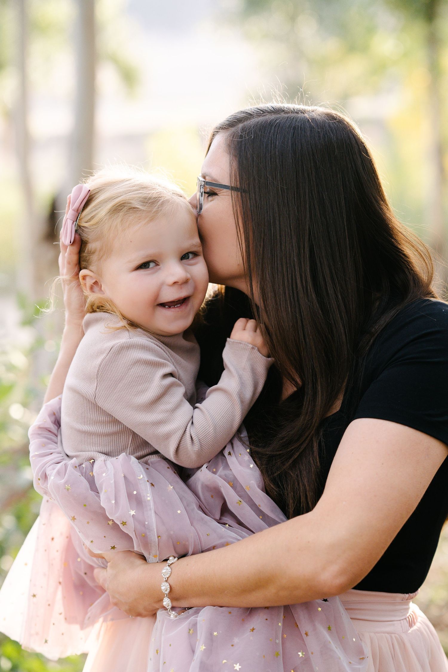 A dark-haired mother kisses her smiling toddler daughter, both wearing pink outfits, in a warm outdoor setting.