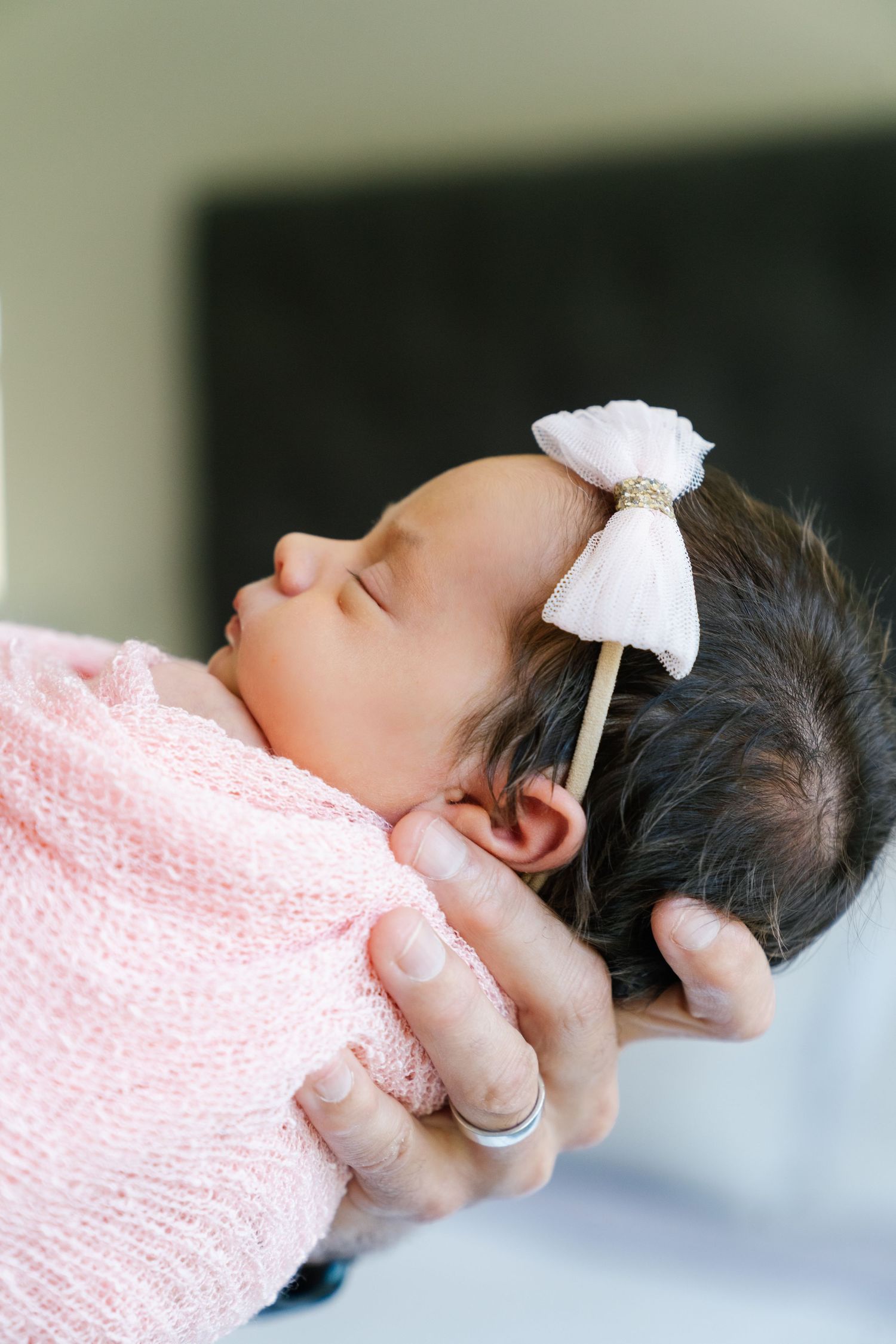 Sleeping infant in pink wrap with white bow accessory held by gentle hands.