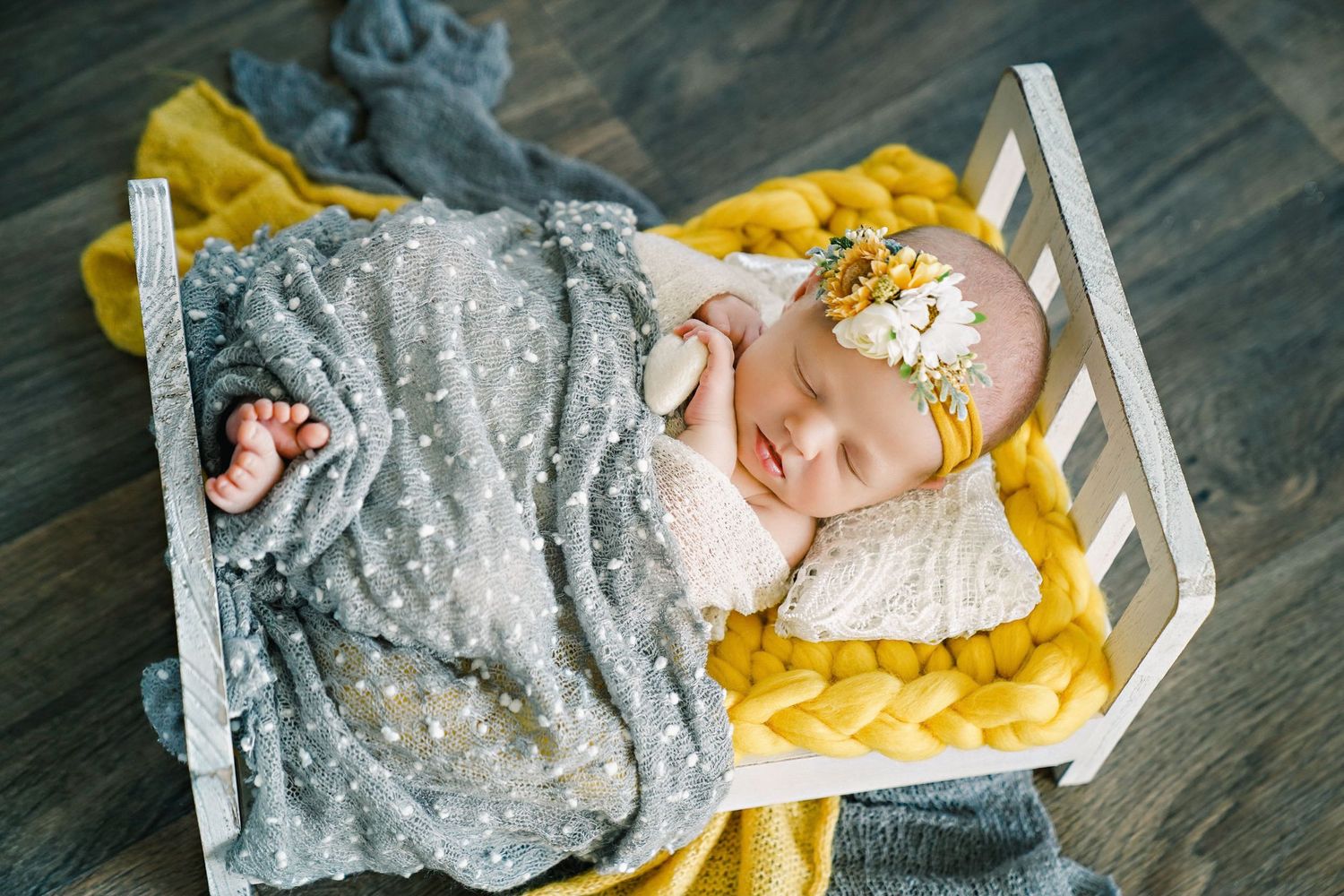 Newborn sleeping in small white bed with gray blanket and yellow bedding.