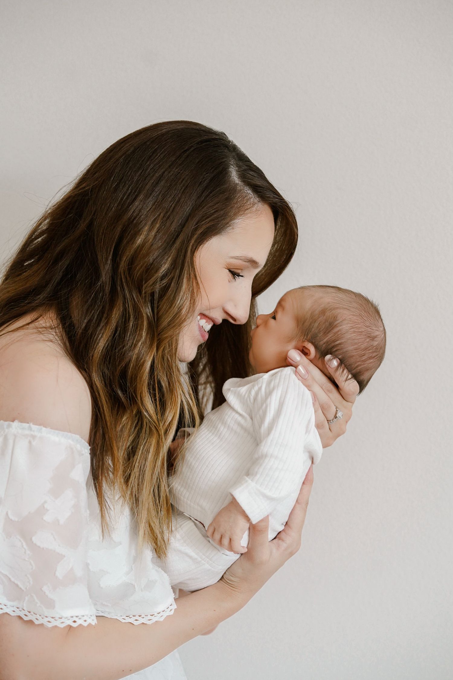 Mother in white lace top holding and gazing at newborn baby against neutral background.