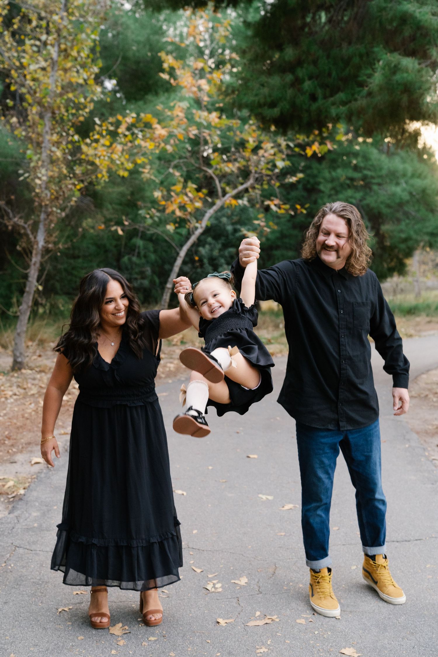 Family plays together on path surrounded by fall foliage, swinging child between them.
