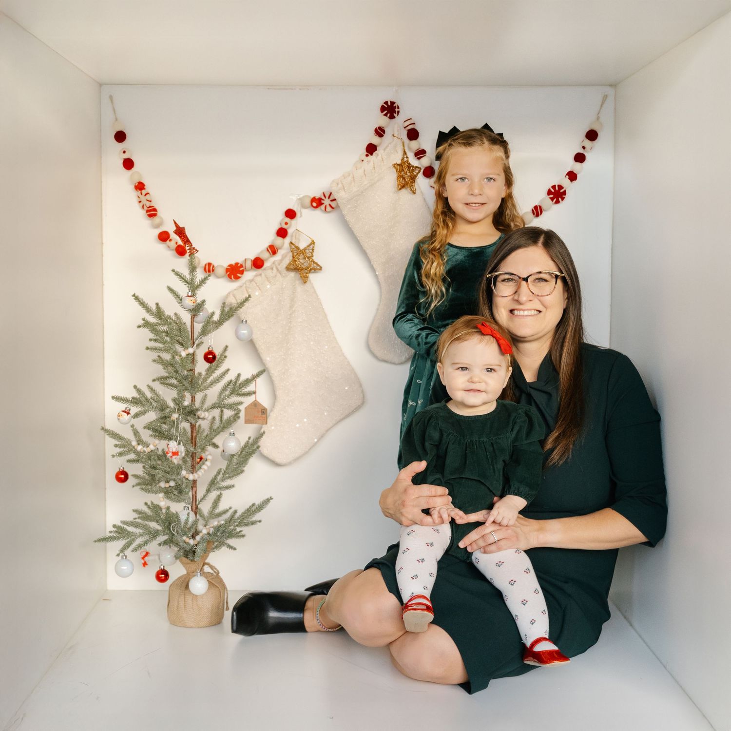 A woman and two young girls pose for a Christmas photo with stockings, garland, and a small decorated tree.
