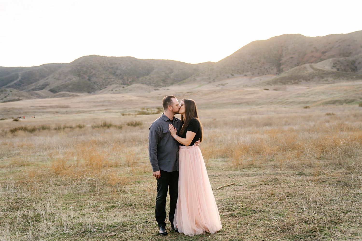 Couple sharing a romantic kiss in a golden field with rolling hills at sunset, woman in pink tulle skirt.