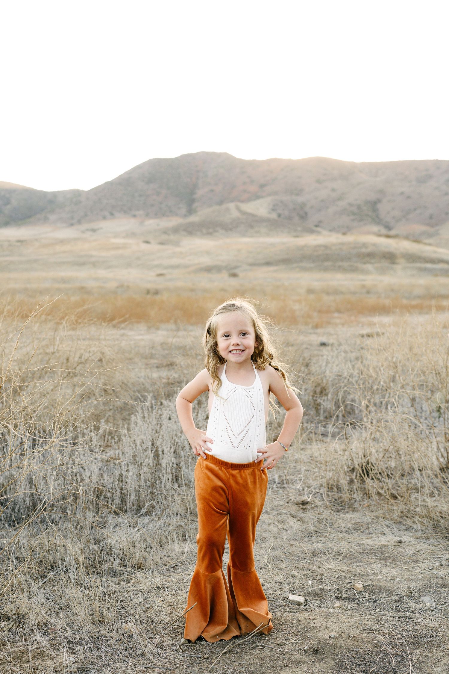 A young girl in rust bell-bottom pants and white top stands confidently in a dry golden meadow with rolling hills.
