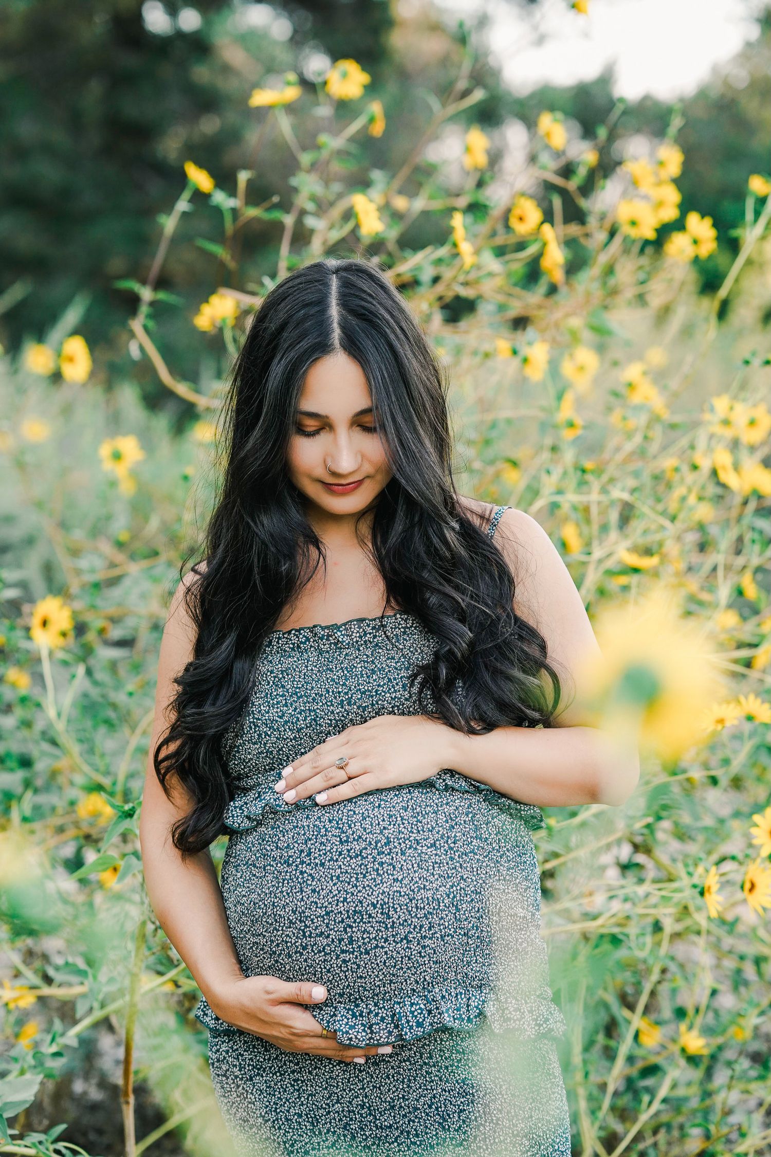 Expectant mother in floral dress caresses baby bump among blooming yellow wildflowers.