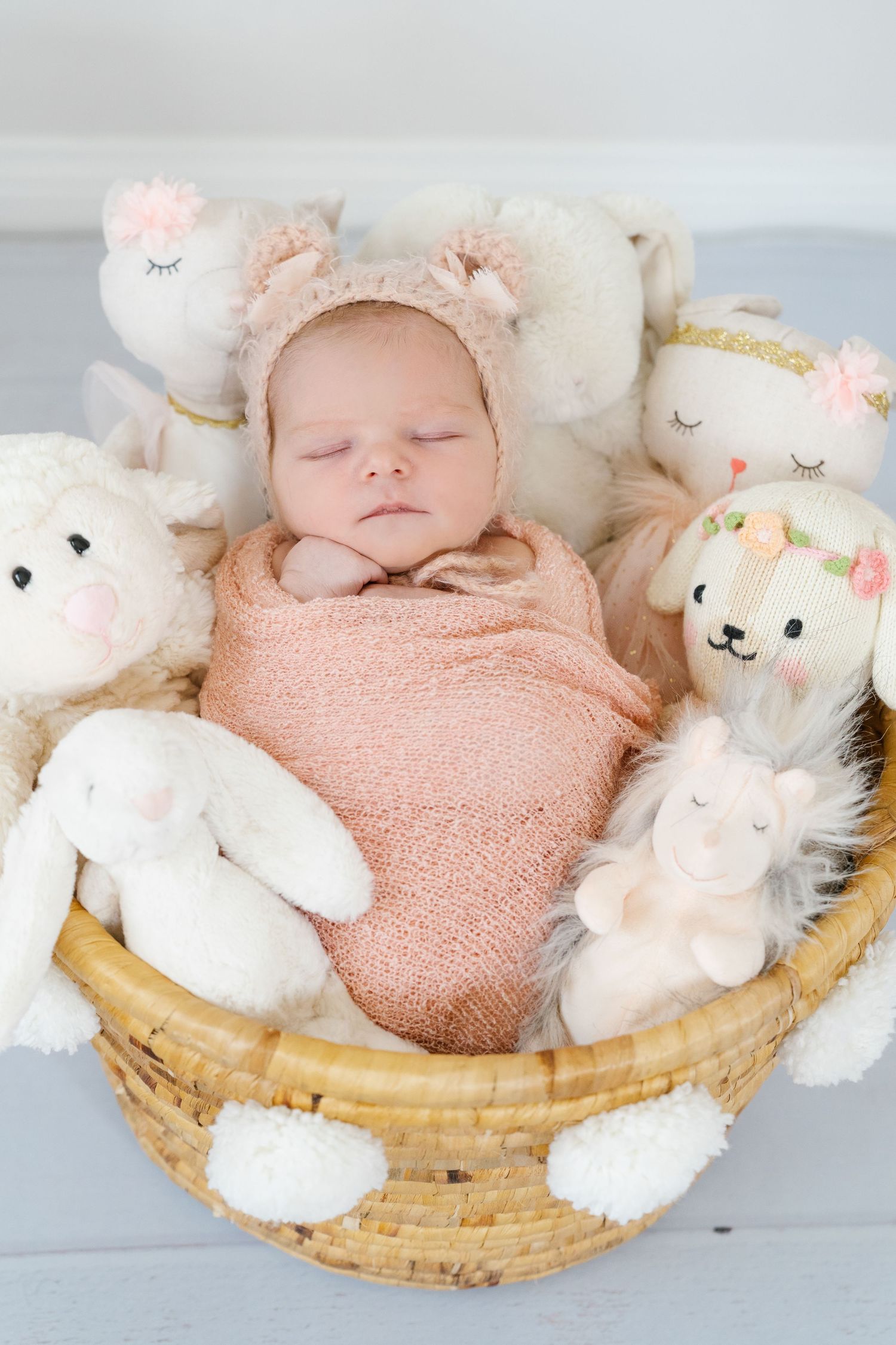 Sleeping baby swaddled in pink surrounded by plush animal toys in a woven basket.