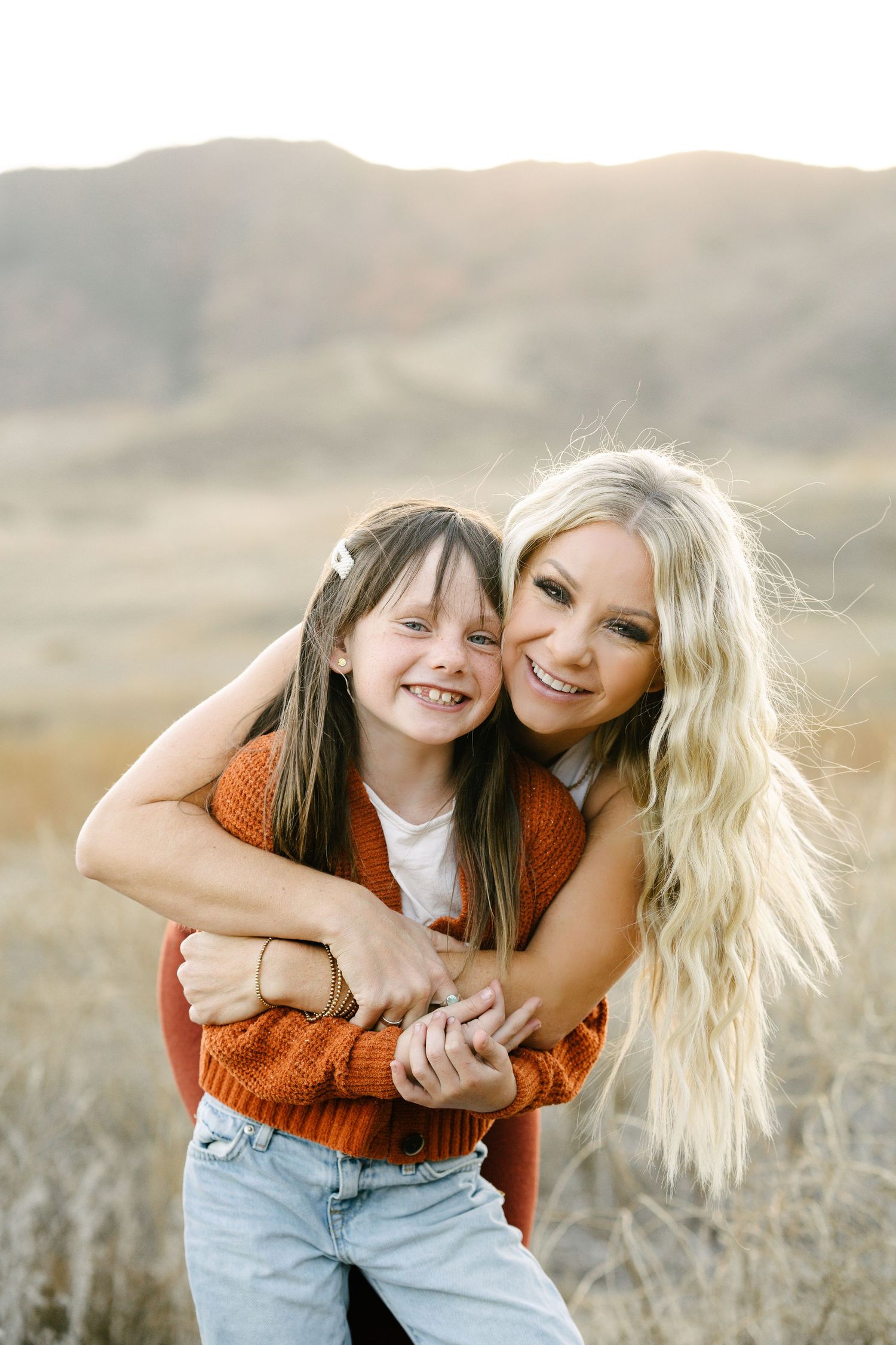 Smiling blonde woman hugging a young girl in an orange sweater outdoors in a golden field.