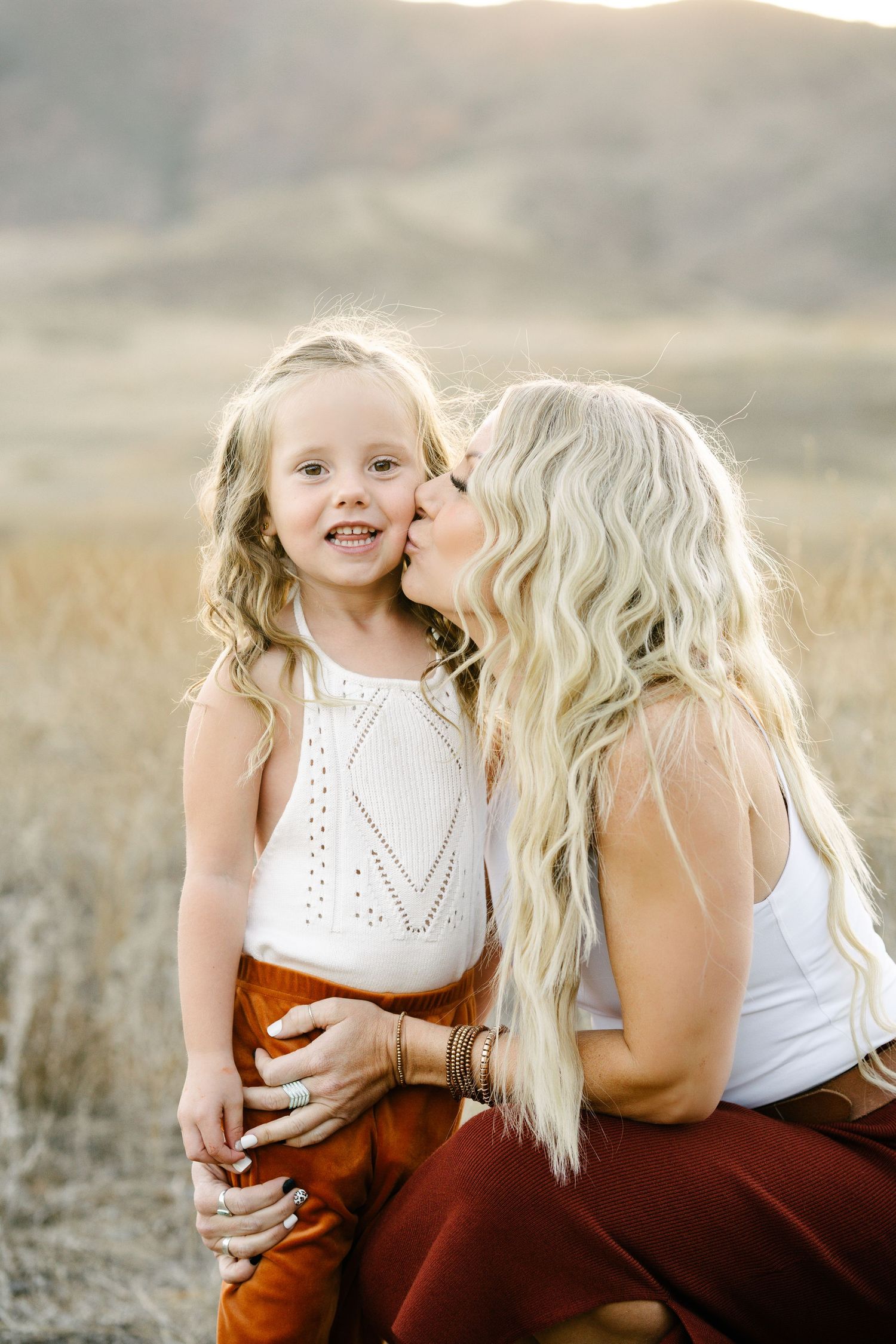 Blonde mother kissing young daughter on cheek in golden field, both wearing white tops and rust-colored bottoms.