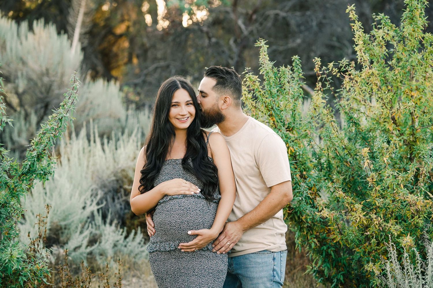 Expecting couple embracing in garden setting with natural desert flora in background during sunset.
